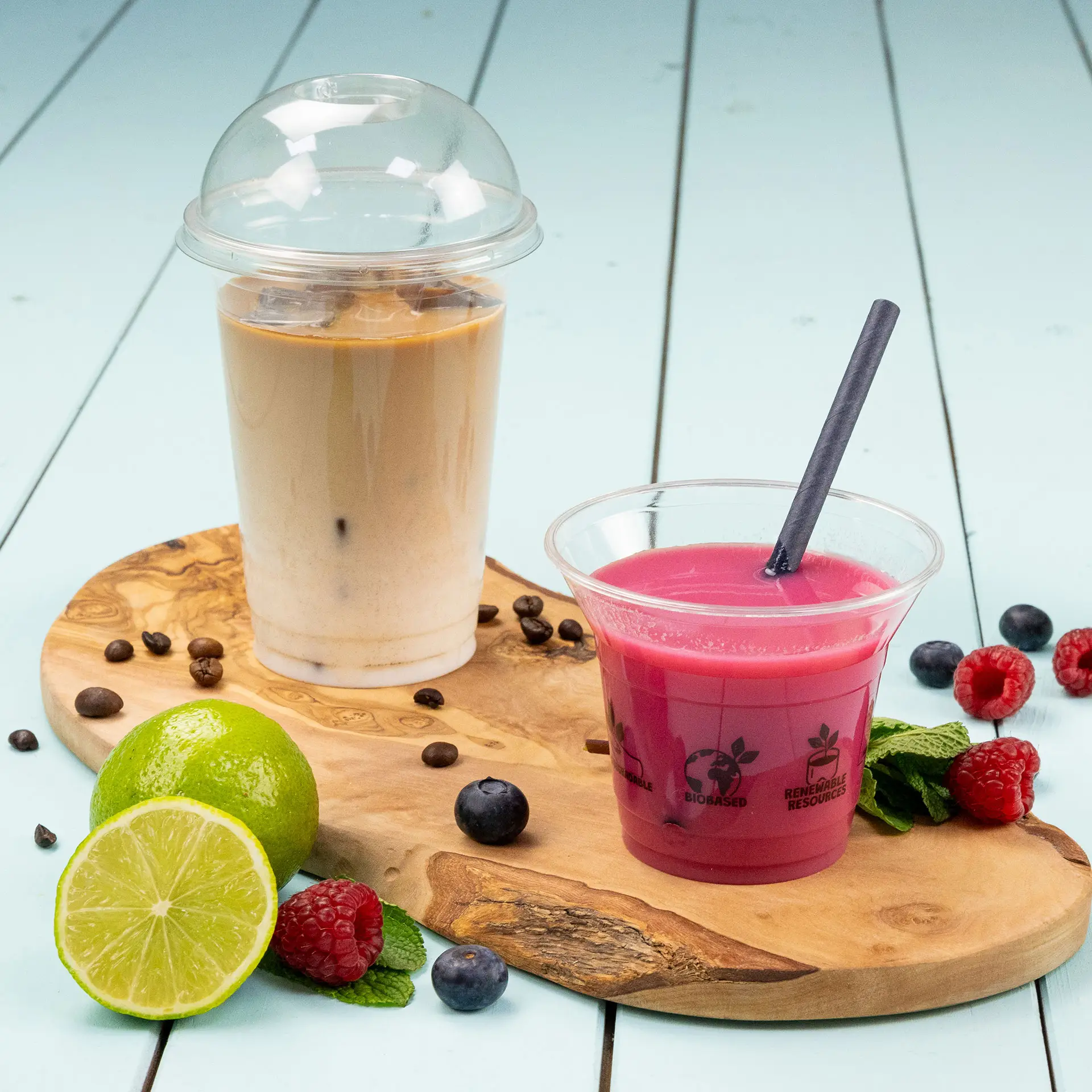 Gobelet en plastique transparent avec du café glacé et une boisson rose, servi sur une planche en bois avec du citron vert et des baies.