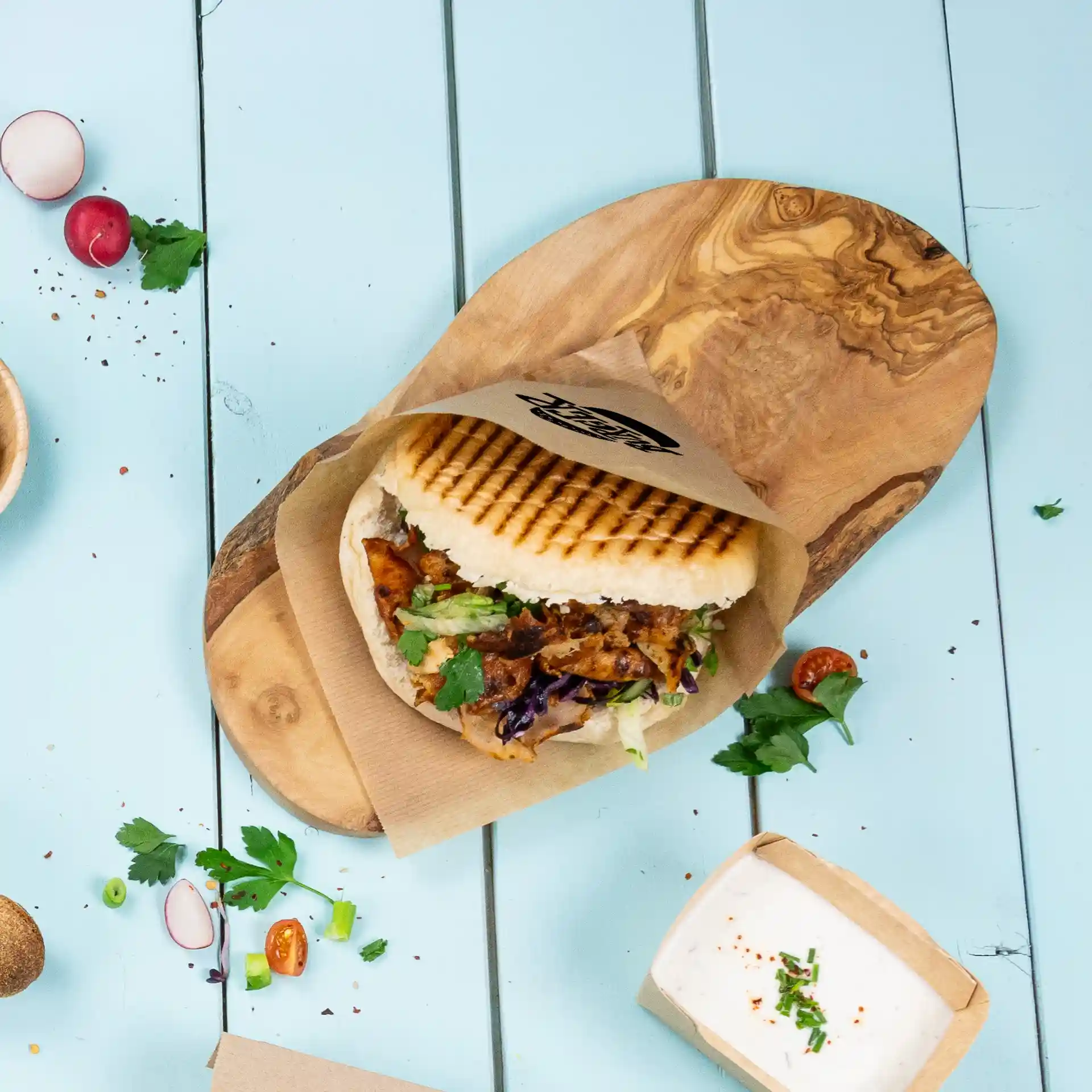 Image d'un sandwich grillé dans une pochette en papier sur une planche en bois, avec des herbes fraîches et un petit contenant de sauce à proximité.