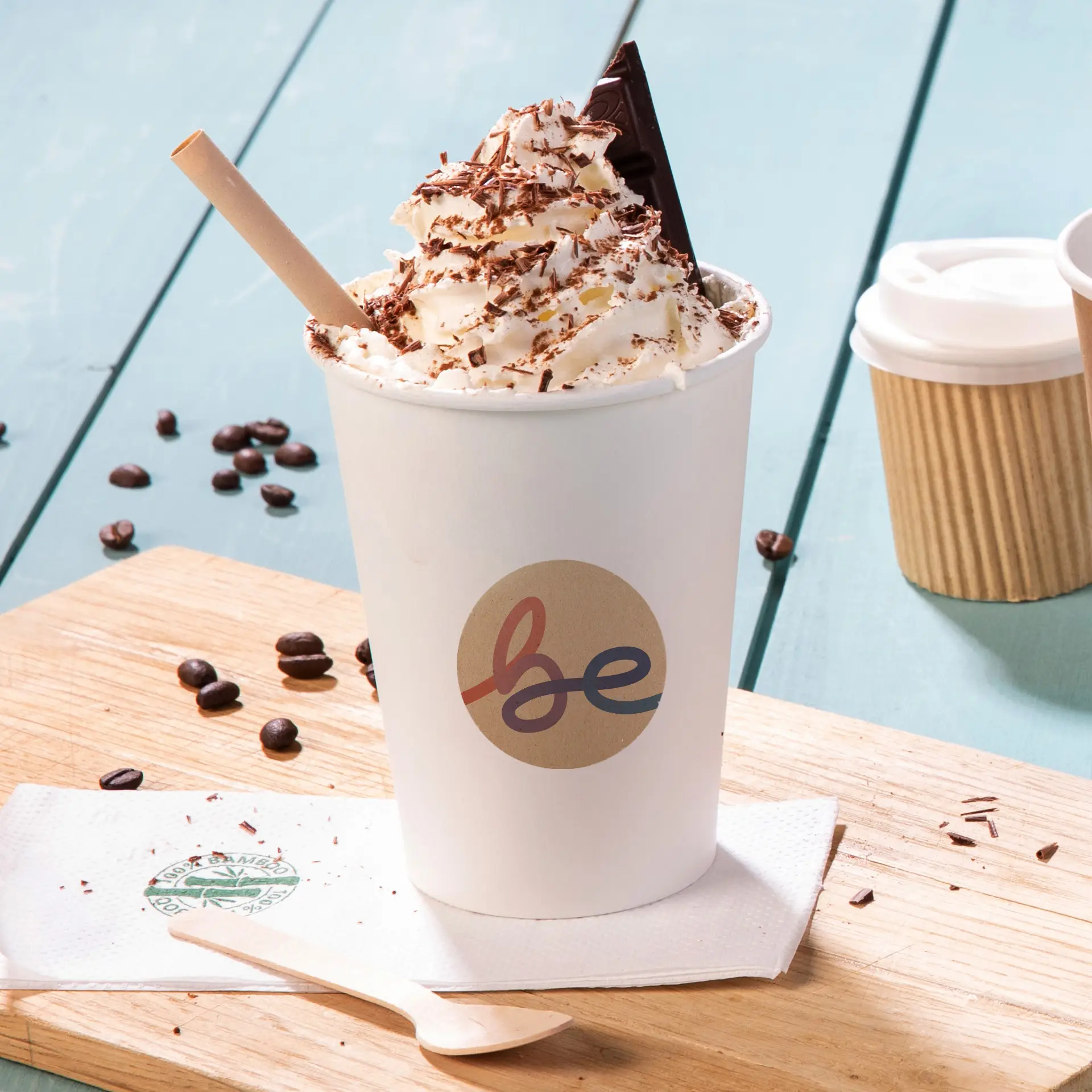 Boisson garnie de crème chantilly dans une tasse blanche avec un logo "be", entourée de grains de café et d'une cuillère en bois.