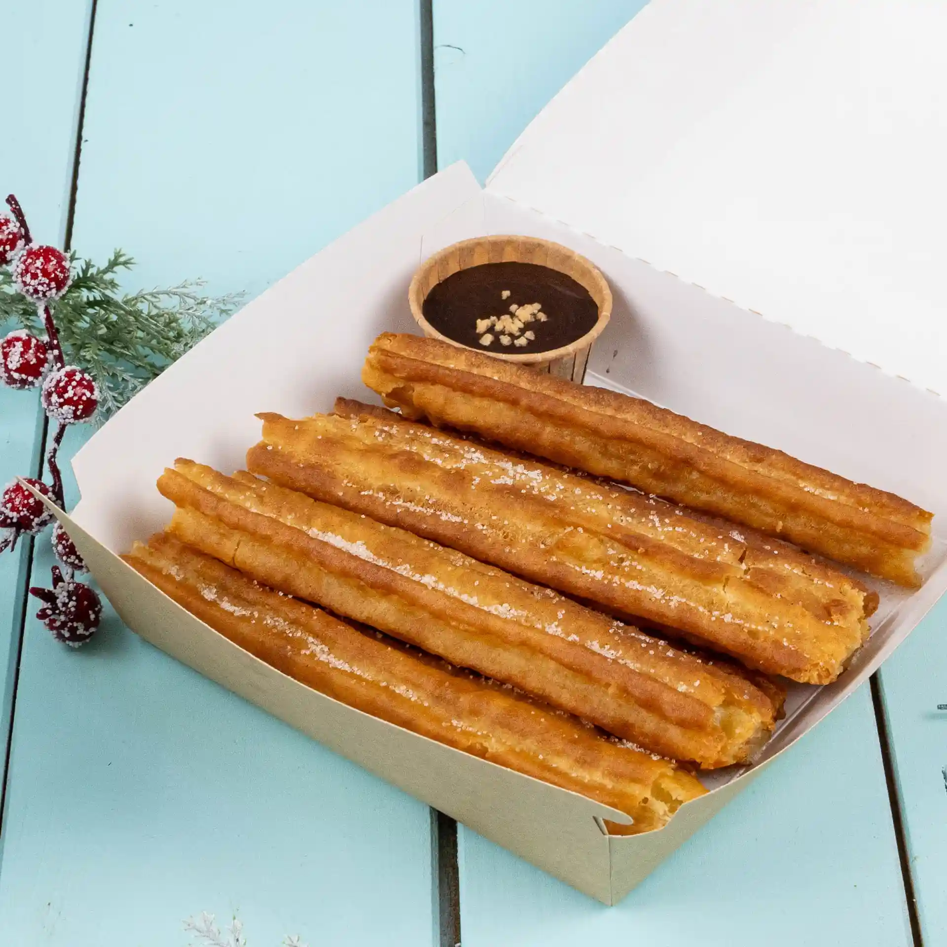Churros dans une boîte en papier avec une petite tasse brune de sauce au chocolat sur une surface bleue.