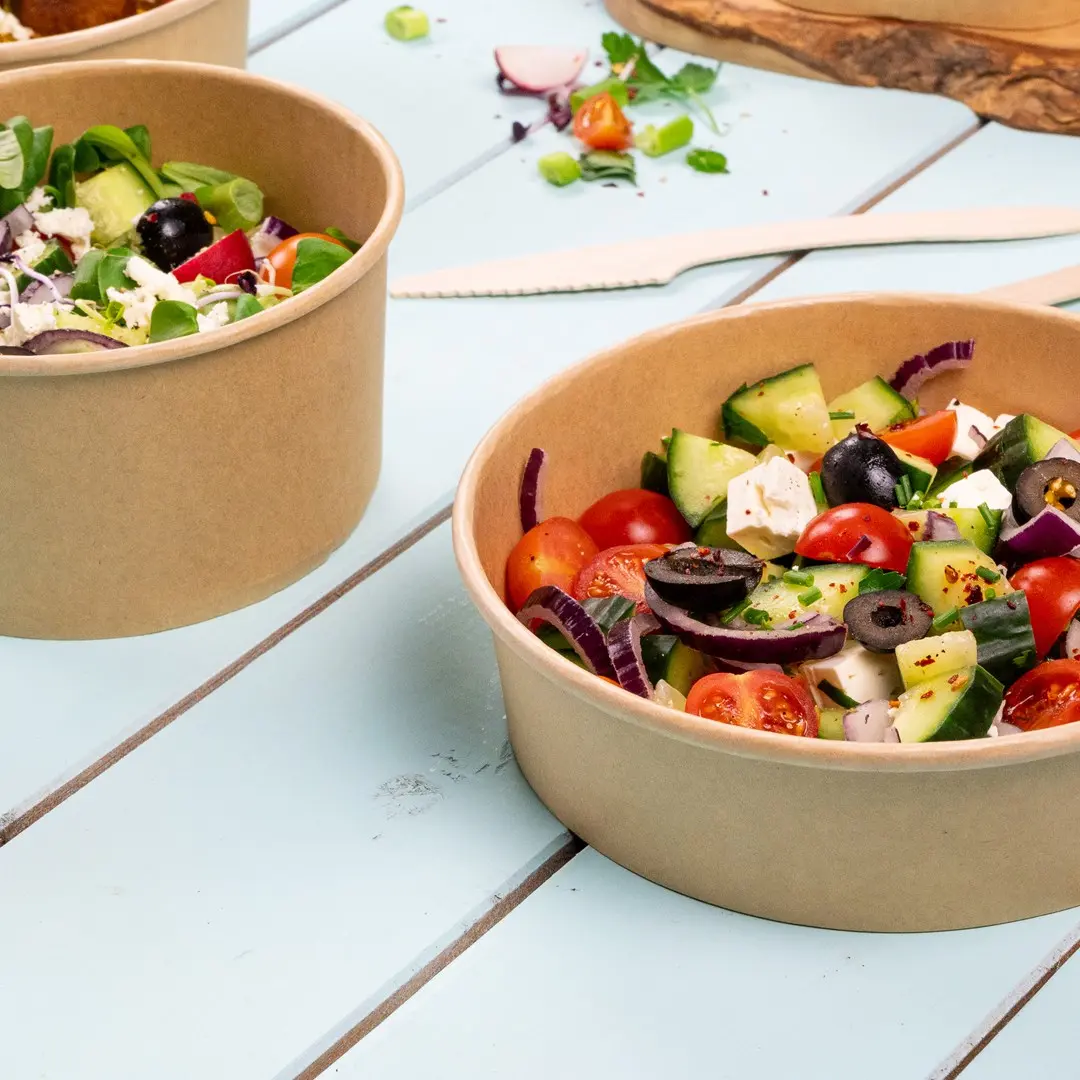 Deux bols en papier marron remplis de légumes mélangés, y compris des tomates, des concombres, des olives et des légumes verts, sur une surface en bois bleu.