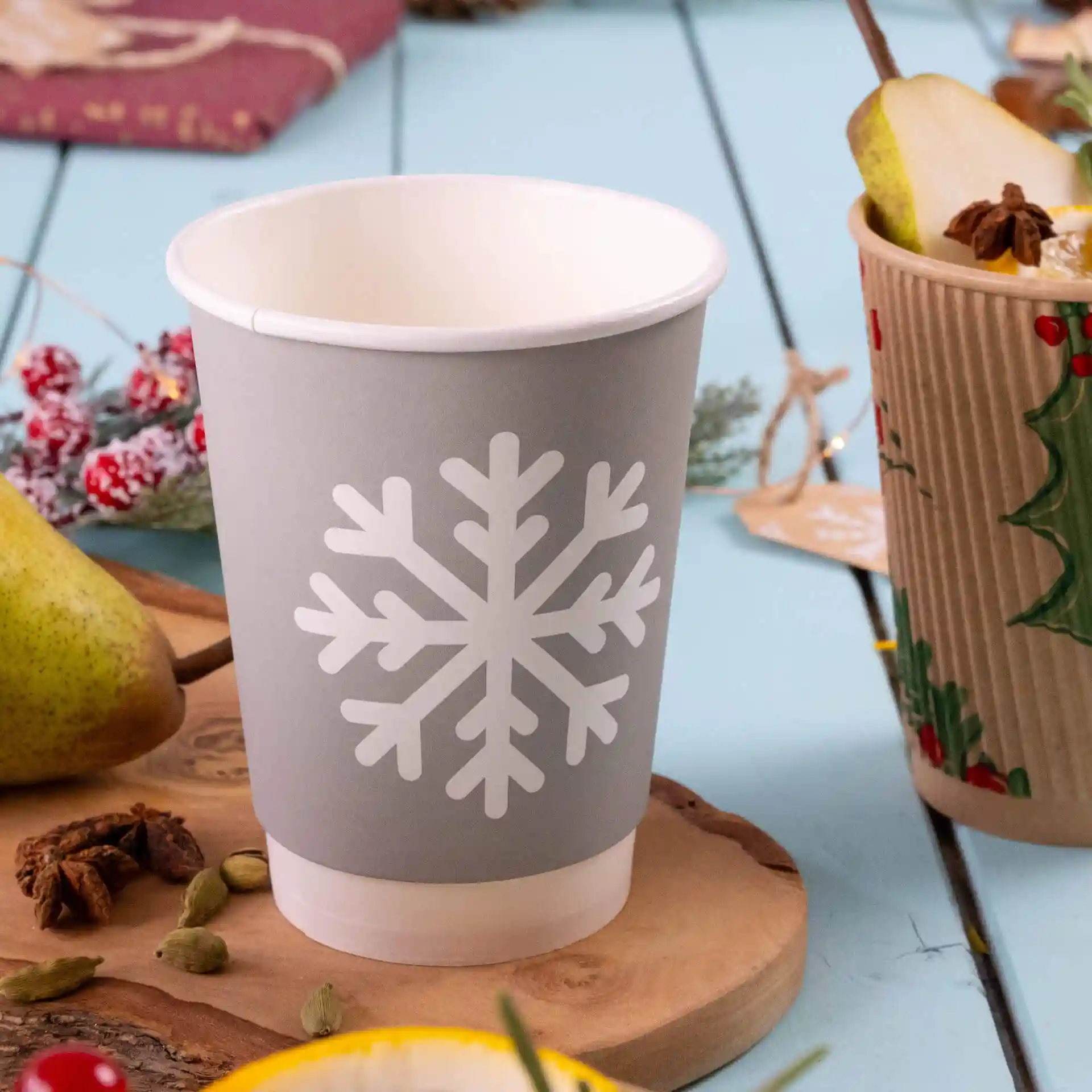 Gobelet en papier gris avec un design de flocon de neige blanc, double paroi, posé sur une surface en bois avec une poire et des épices à proximité.