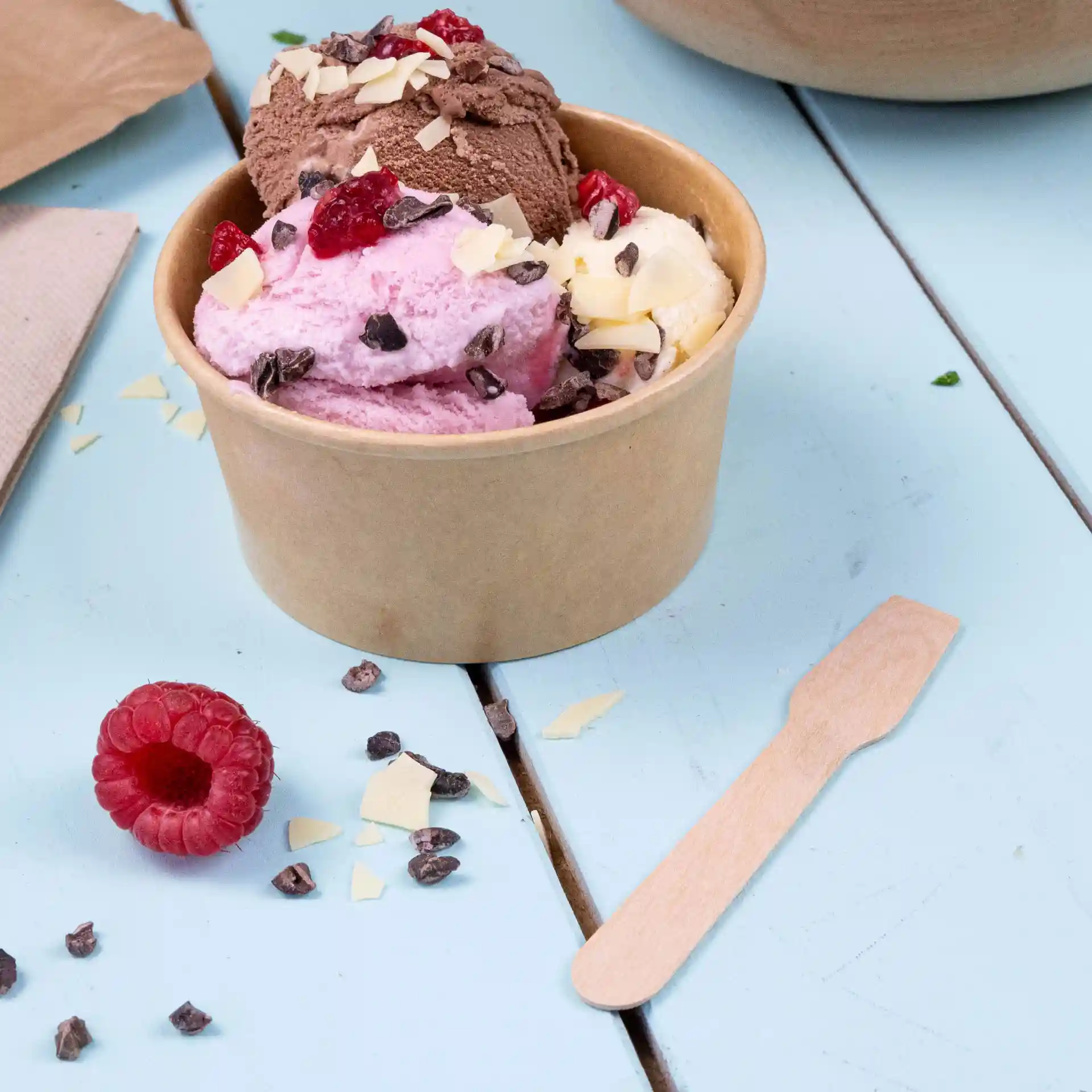 Gobelet contenant trois boules de glace rose, brune et jaune, surmonté de pépites de chocolat et de framboises, avec une cuillère en bois à côté.