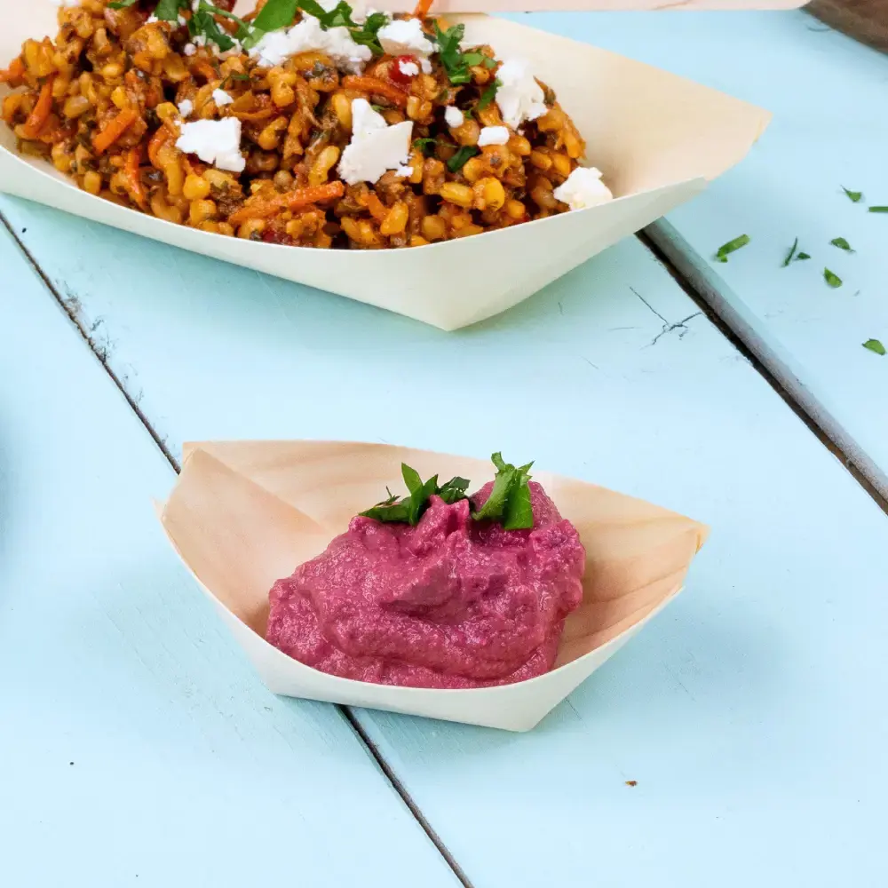 Plat en forme de bateau en bois contenant une sauce rose garnie d'herbes vertes, à côté d'un plat plus grand de grains et légumes mélangés.