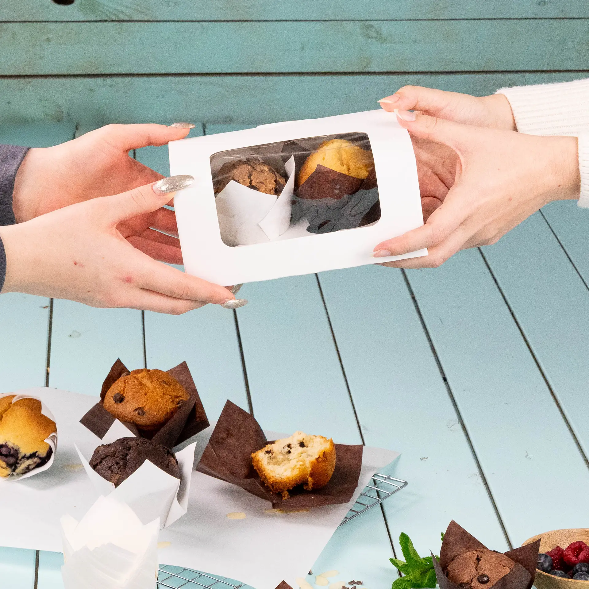 Des mains échangent une boîte transparente contenant deux muffins, avec plusieurs autres muffins dans des cups en papier brun sur une table.