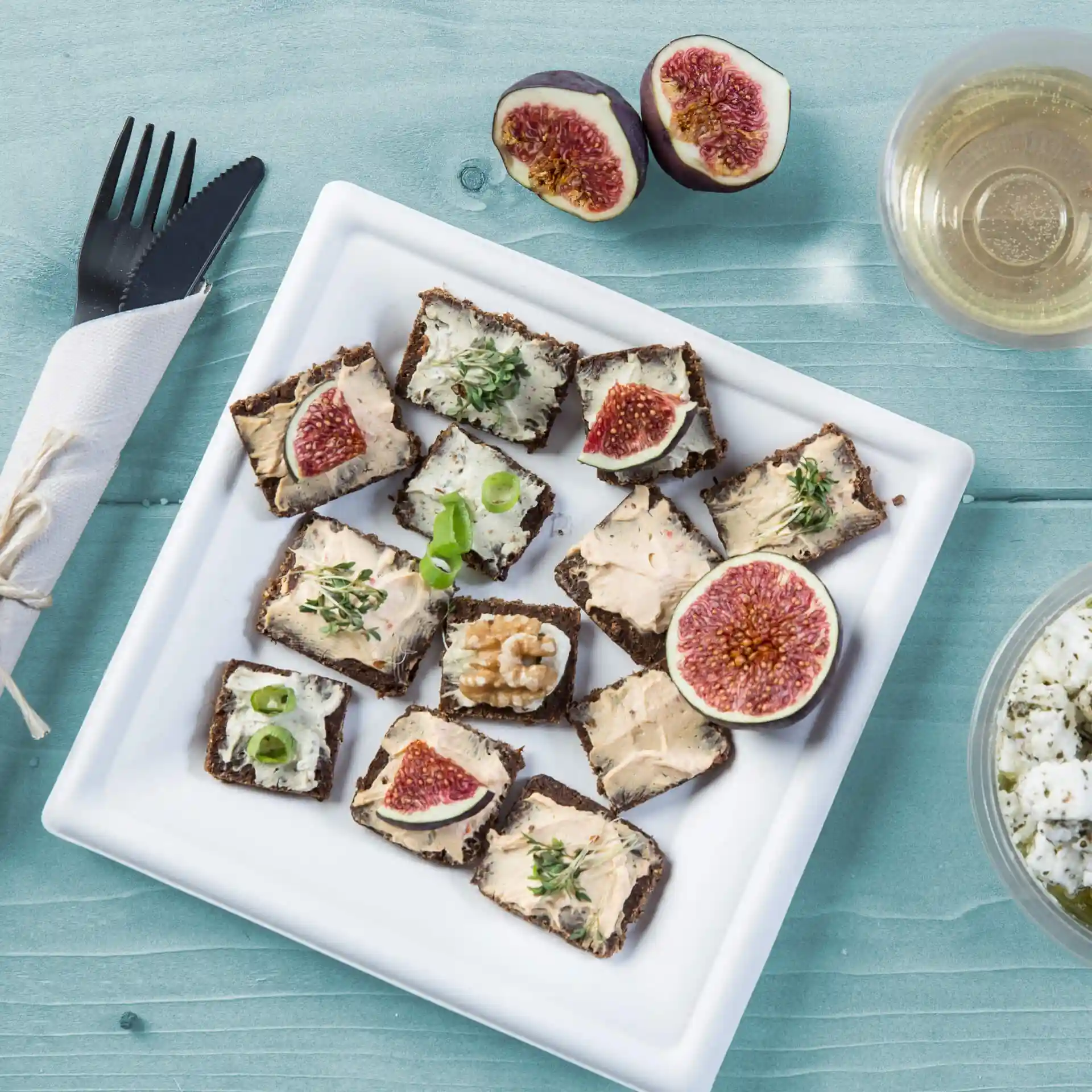 Assiette avec des assortiments d'apéritifs, y compris du pain garni de tartinades, des figues et des herbes, sur une assiette carrée en canne à sucre blanche.