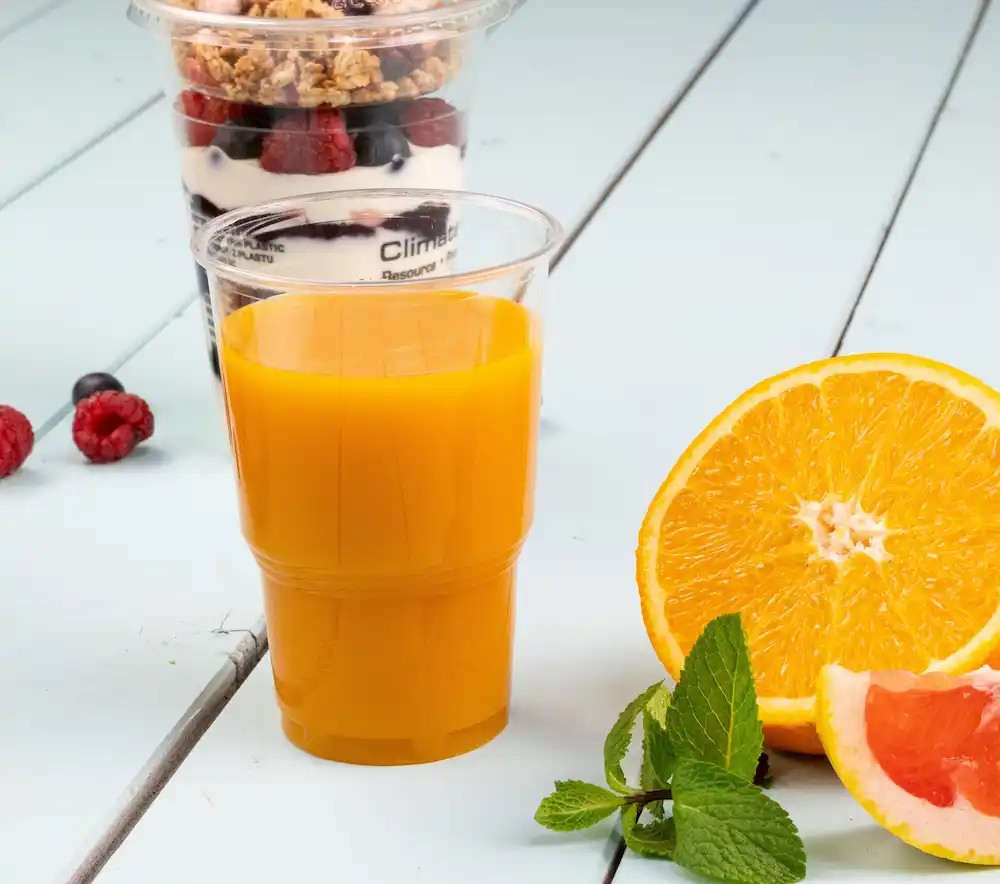 Boisson orange dans un verre transparent à côté d'une orange coupée en deux et de feuilles de menthe, avec un verre à dessert en arrière-plan.