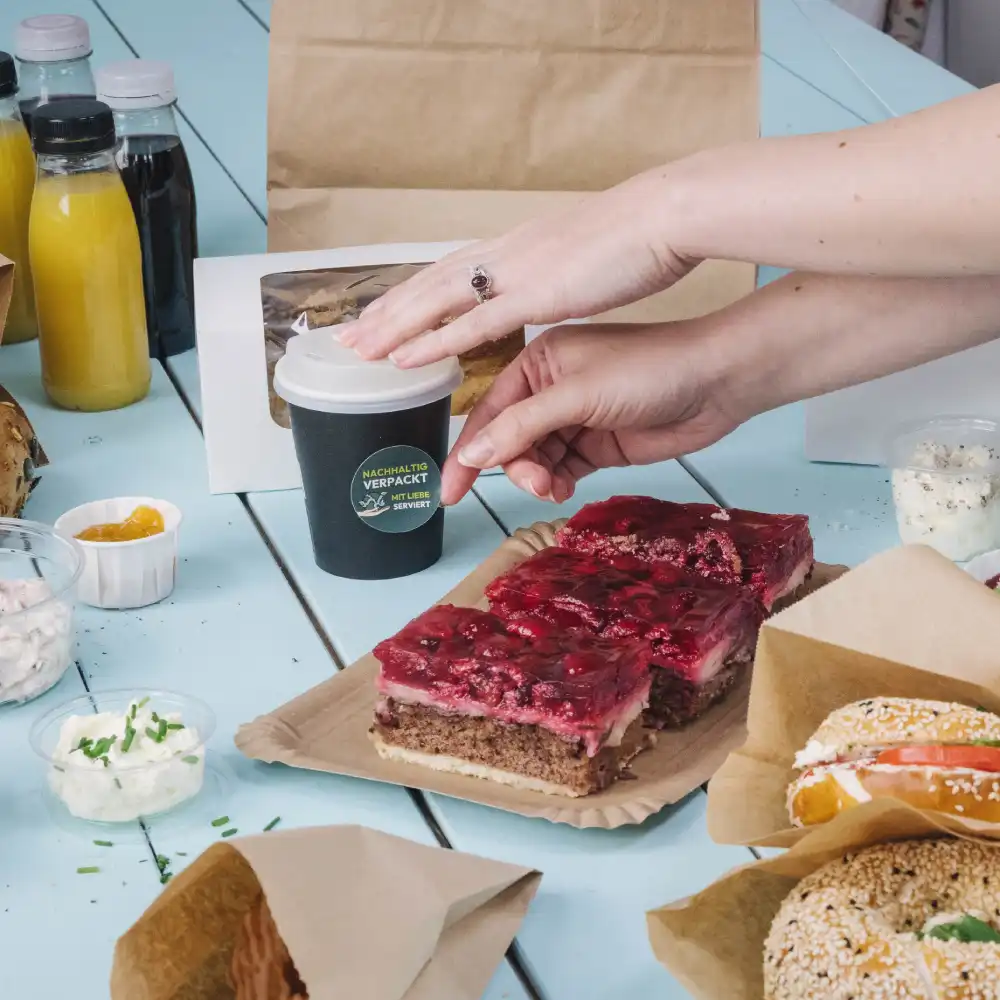 Une main atteignant une petite tasse de café noire sur une table avec divers aliments ; gâteau en couches, sauces et boissons à proximité.