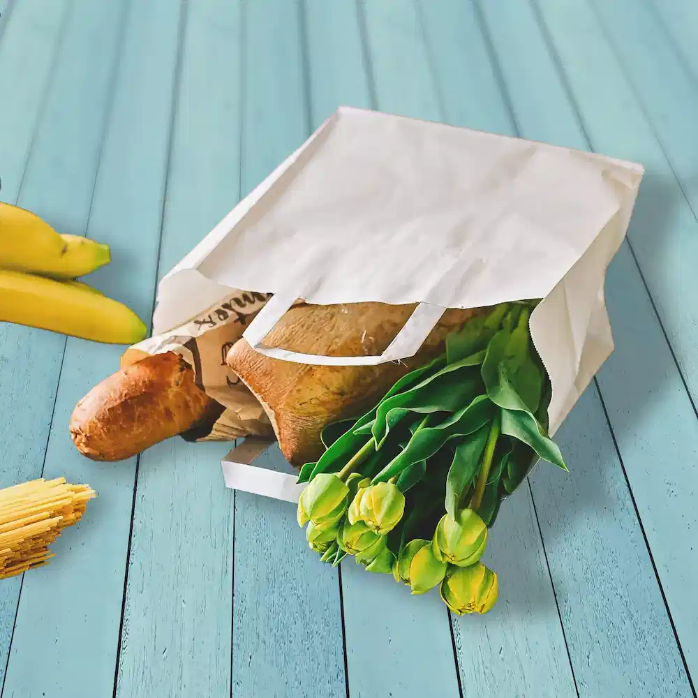 Sac en papier blanc rempli de courses, y compris du pain, des tulipes et des bananes sur une surface en bois bleu.