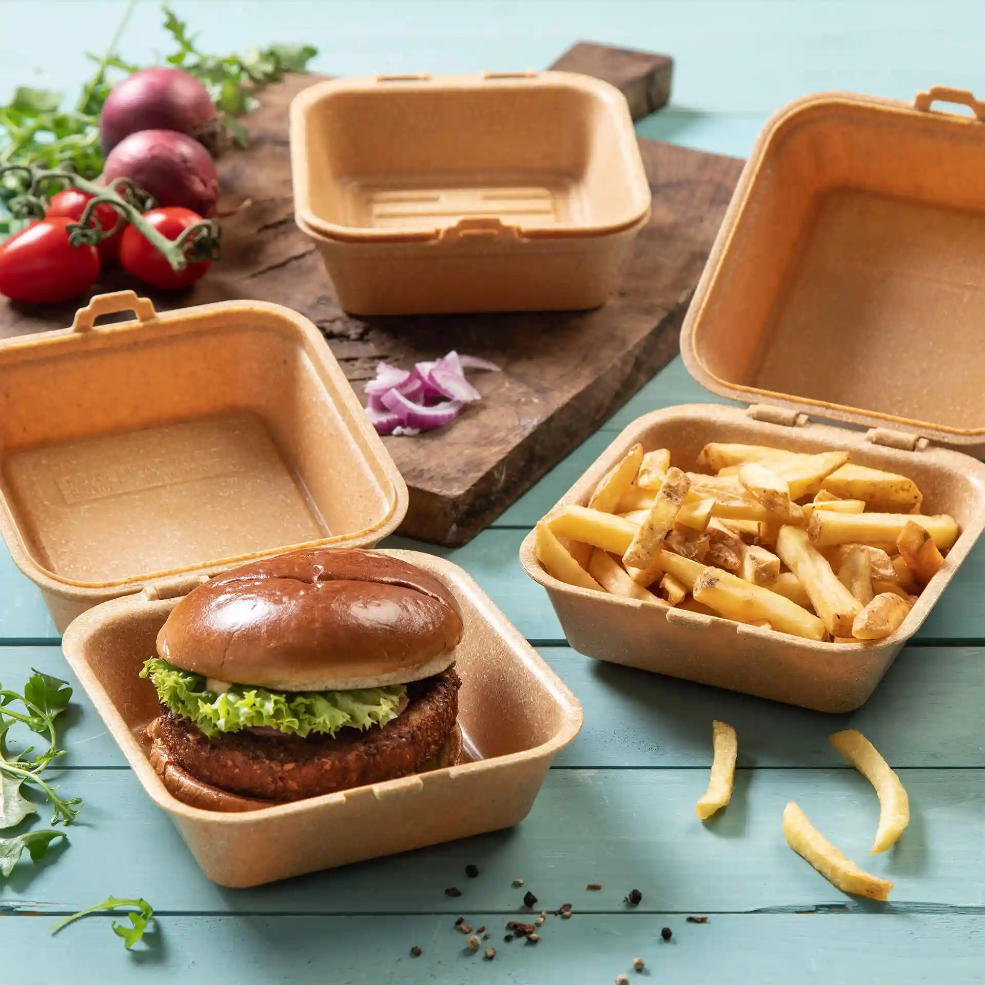 Boîte à burger réutilisable marron avec couvercle, contenant un burger et des frites, sur une surface en bois avec des légumes à proximité.