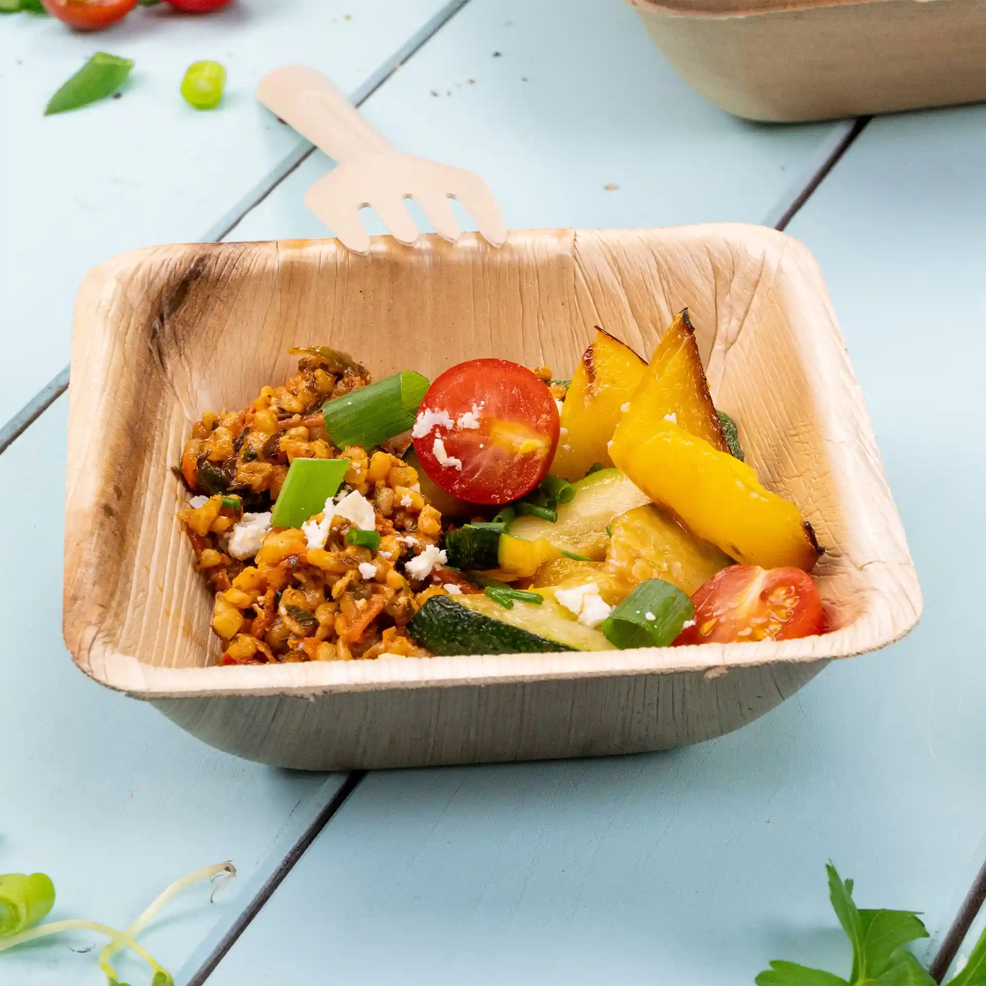 Bol carré en feuille de palmier rempli de riz, légumes et une tomate cerise, avec une fourchette en bois reposant sur le bord.