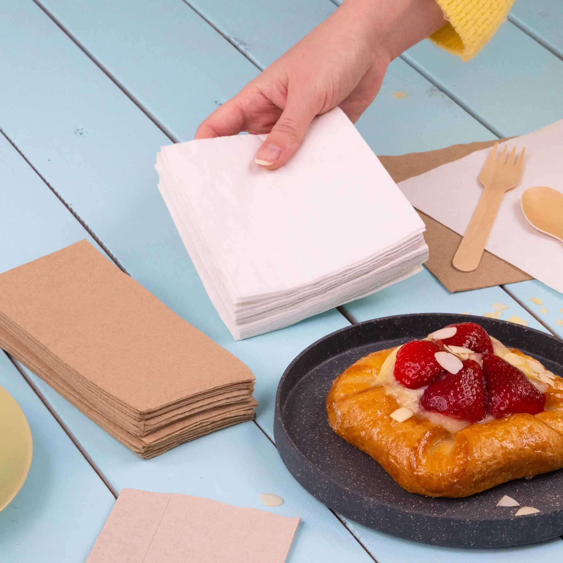 Serviettes à cocktail blanches en papier 2 plis empilées dans une main, avec un croissant garni de fraises sur une assiette à côté.