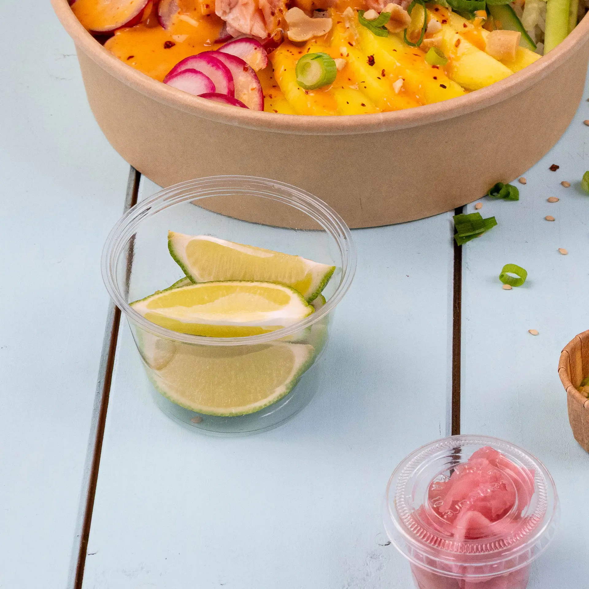 Coupes en PLA transparentes contenant des quartiers de lime et des oignons roses marinés, placées à côté d'un bol d'ingrédients de salade.