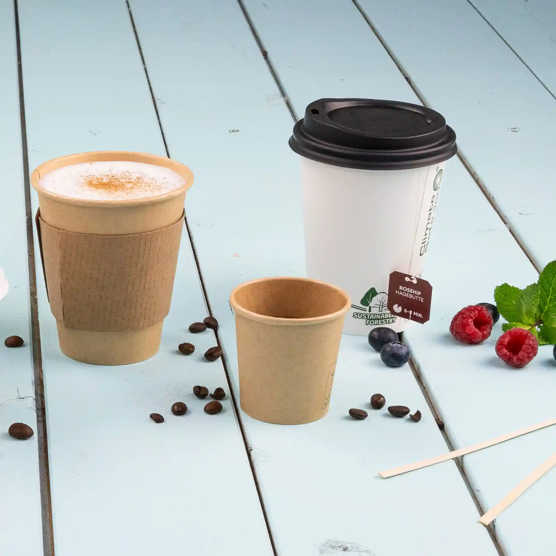 Image de trois gobelets en papier sur une surface en bois bleu, y compris un gobelet blanc de 300 ml avec un couvercle noir, un gobelet marron et un plus petit gobelet marron.