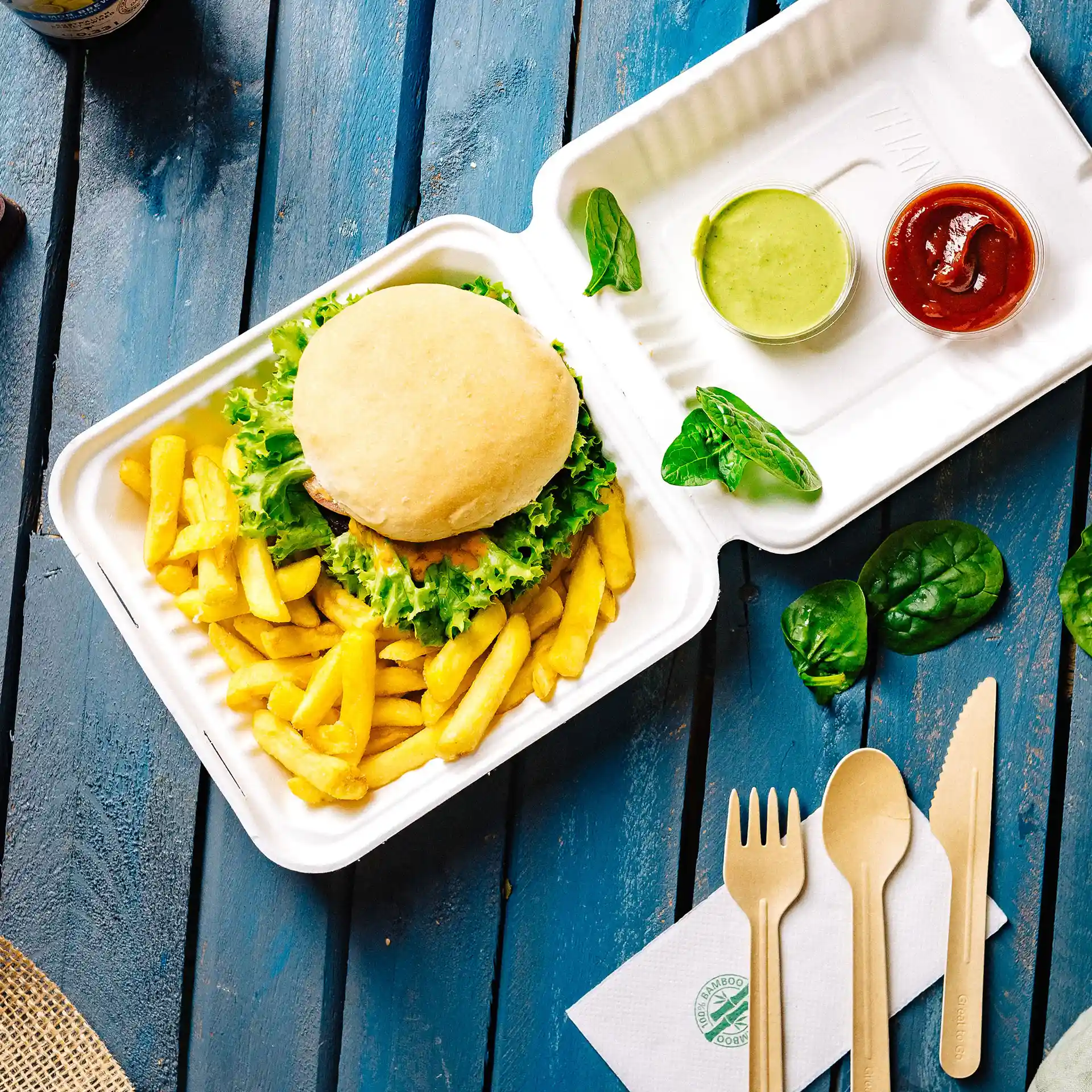 Conteneur rectangulaire blanc avec couvercle, contenant un burger, des frites, une sauce verte, du ketchup et des ustensiles en bois sur une surface bleue.