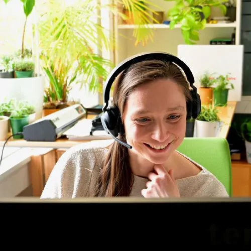 Femme avec des écouteurs souriante assise à un bureau avec des plantes et un ordinateur dans une pièce lumineuse.