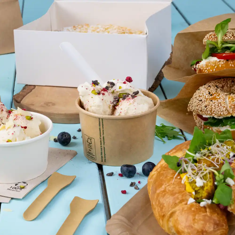 Crème glacée dans un verre avec des garnitures, deux sandwiches sur du papier, un croissant garni et des myrtilles sur une surface en bois bleue.
