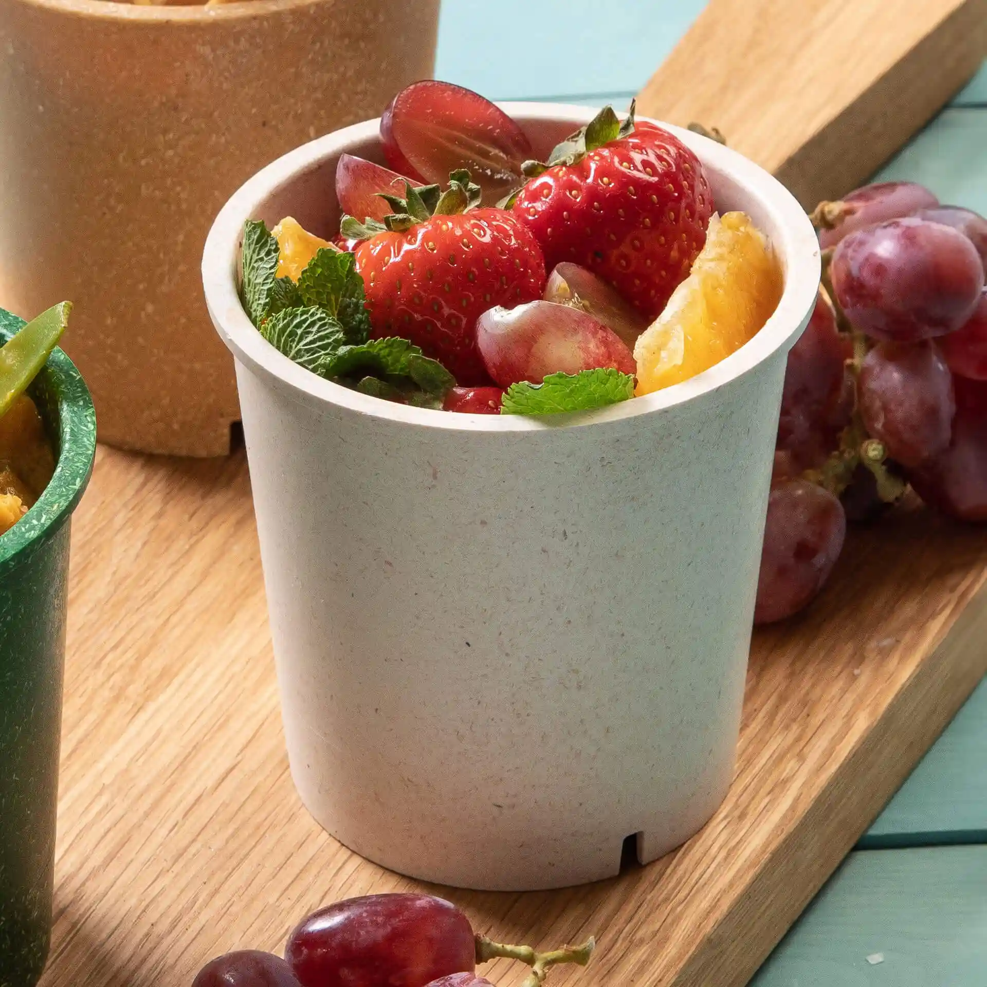 Gobelet rond réutilisable crème rempli de fraises, raisins et feuilles de menthe sur une surface en bois.