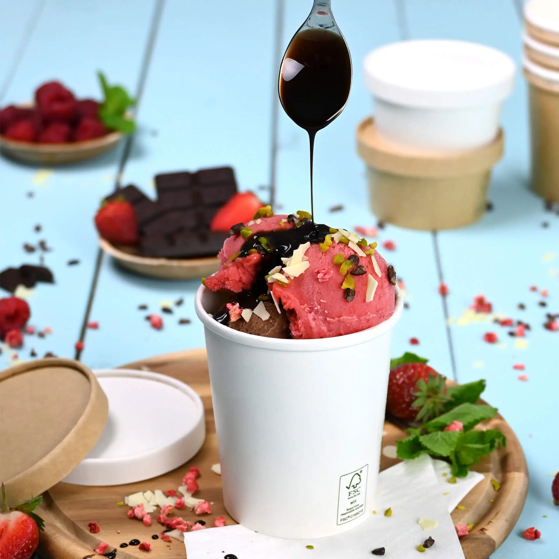 Un gobelet blanc de glace rempli de glace au chocolat et à la fraise, garni de sauce au chocolat, de pistaches et de chocolat râpé, sur un plateau en bois entouré de fraises fraîches, de framboises et de carrés de chocolat.