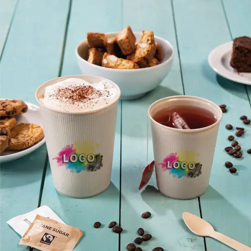 Deux tasses avec design de logo, l'une avec de la crème fouettée et l'autre avec du thé glacé, entourées de biscuits, de gâteau et de sachets de sucre.