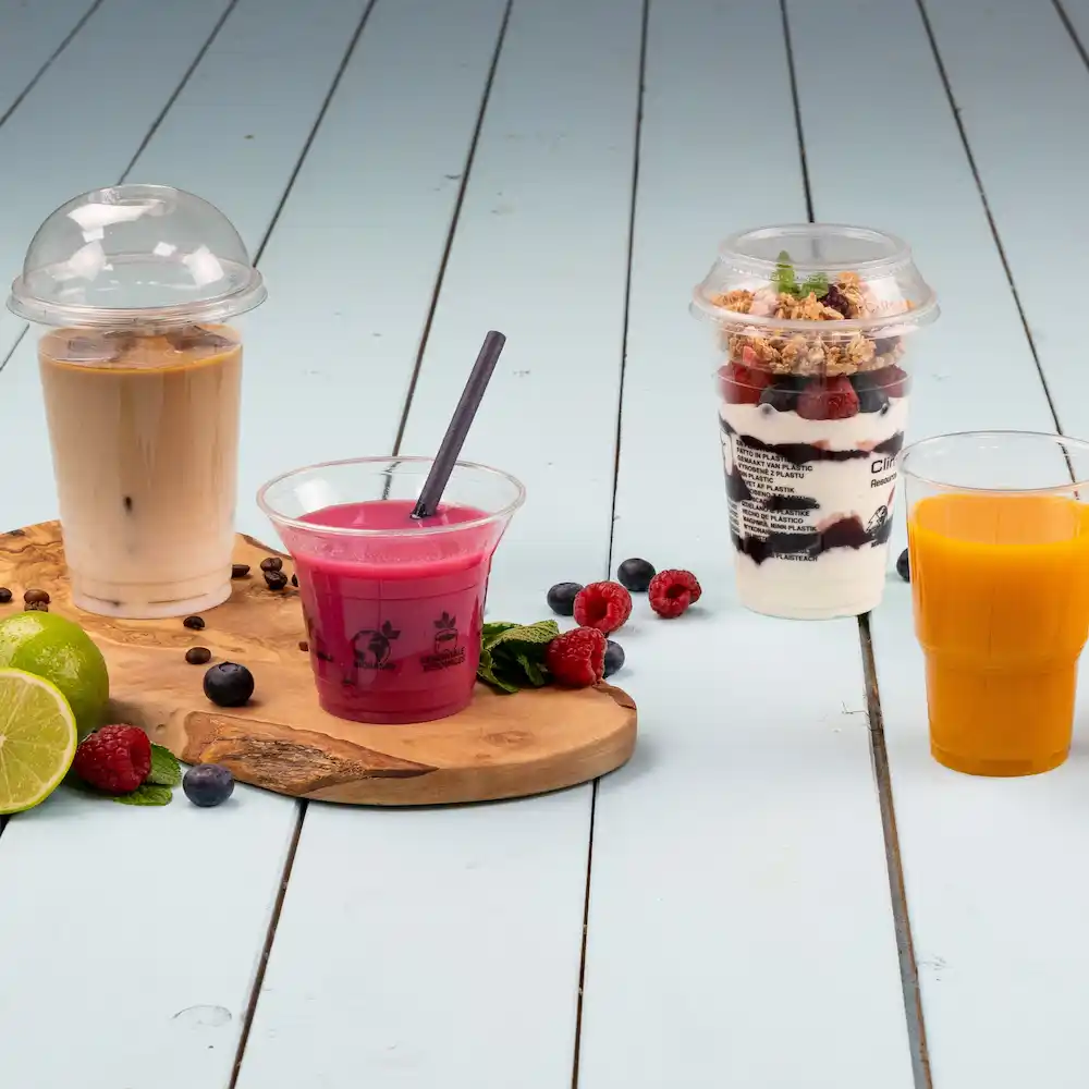 Quatre boissons sur une planche en bois : café glacé dans un gobelet avec un couvercle en dôme, boisson rose avec une paille, verre à dessert en couches et jus d'orange.