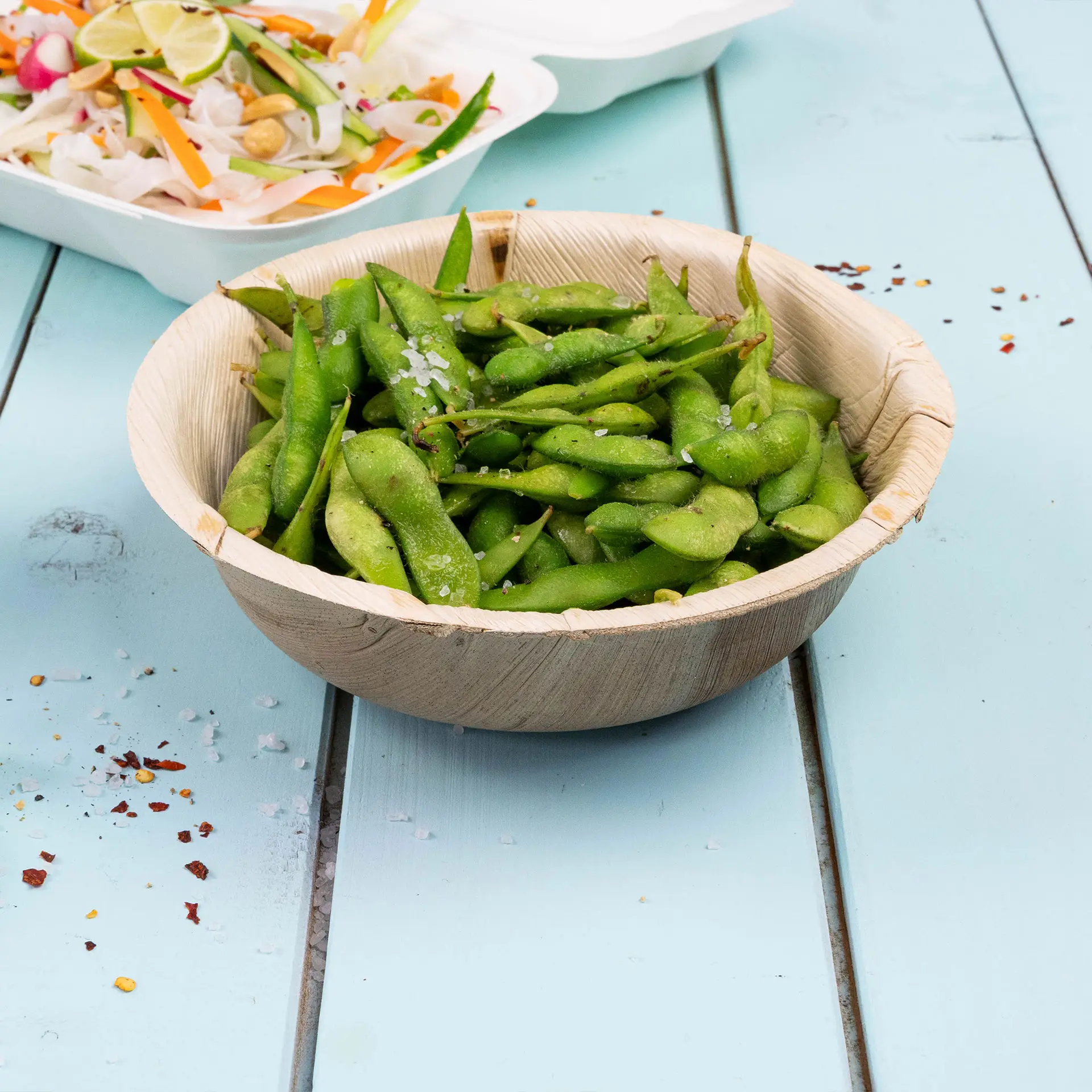Bol en feuille de palmier rond rempli d'edamame vert, saupoudré de sel, sur une surface en bois bleu.