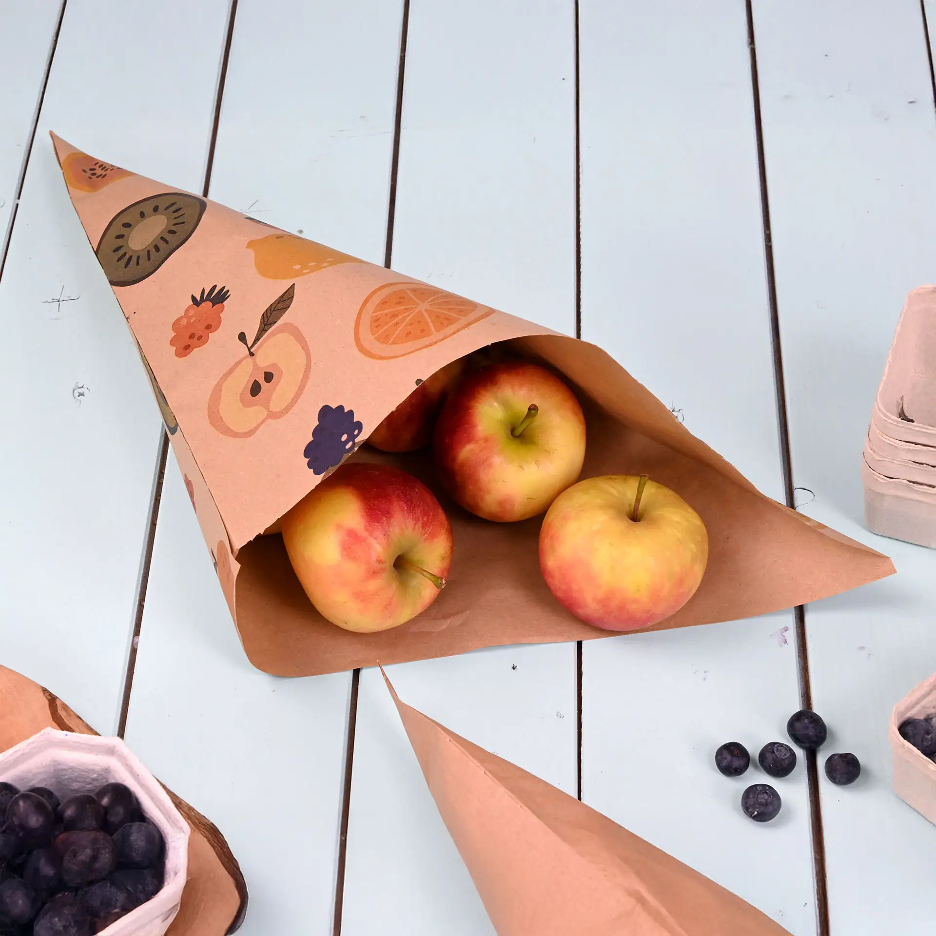Cone en papier brun avec un motif de fruits, contenant trois pommes rouges, sur une surface de table bleu clair.