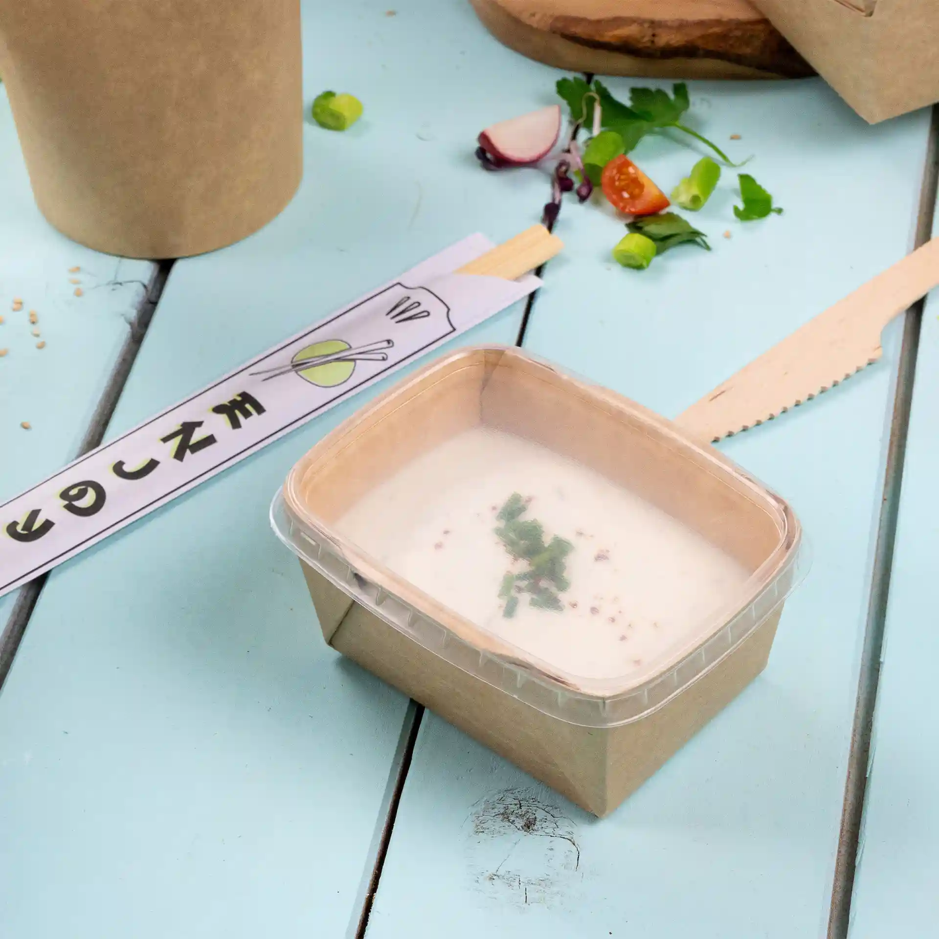Conteneur rectangulaire transparent avec couvercle, contenant une soupe crémeuse garnie d'herbes, sur une surface bleu clair.