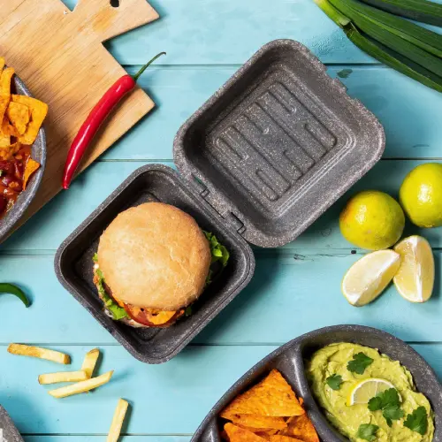 Burger dans une boîte kraft réutilisable avec de la laitue et des sauces, entouré de chips, frites, guacamole et quartiers de lime.