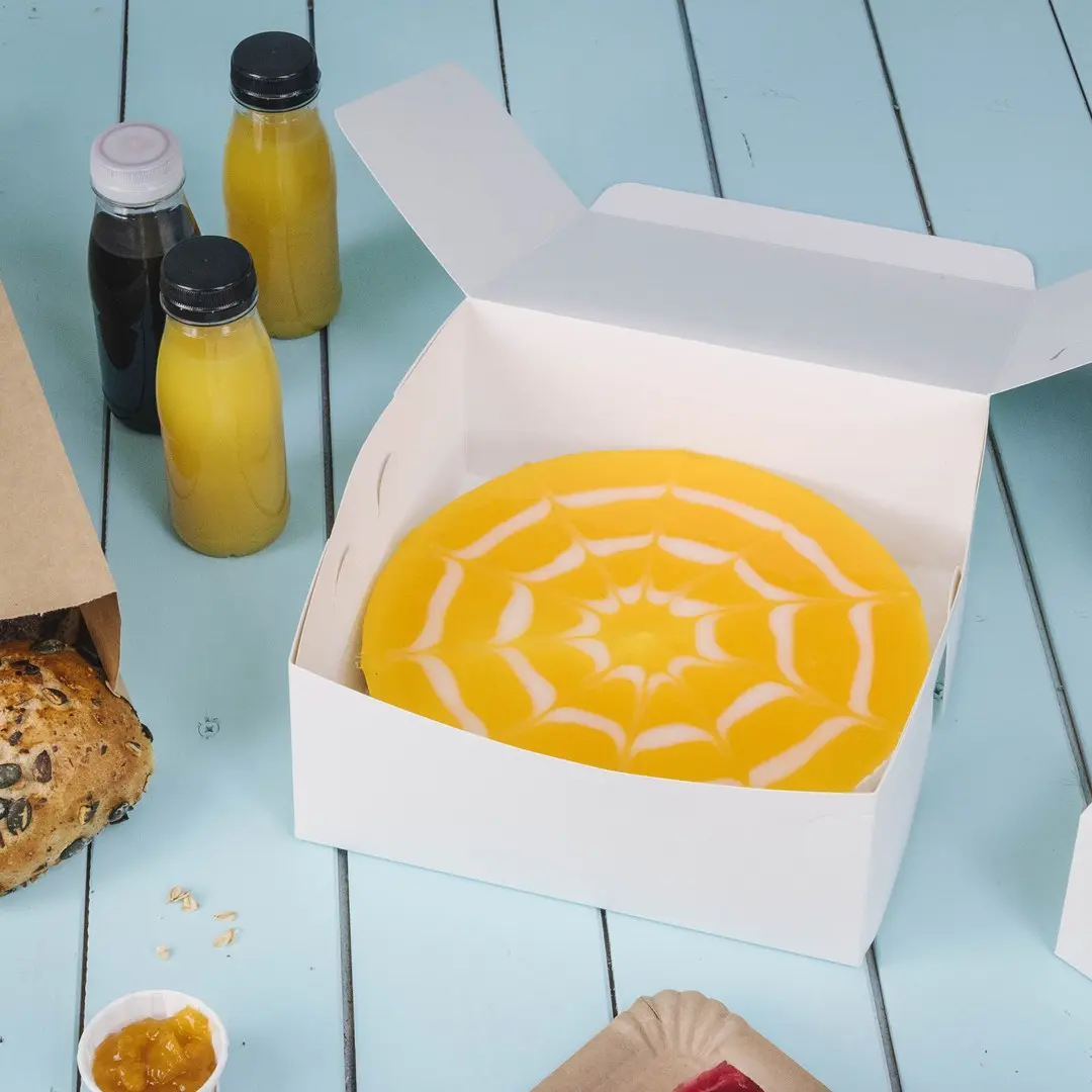 Boîte à gâteau blanche contenant un gâteau jaune avec un design de toile d'araignée blanche, entourée de boissons et de pâtisseries.