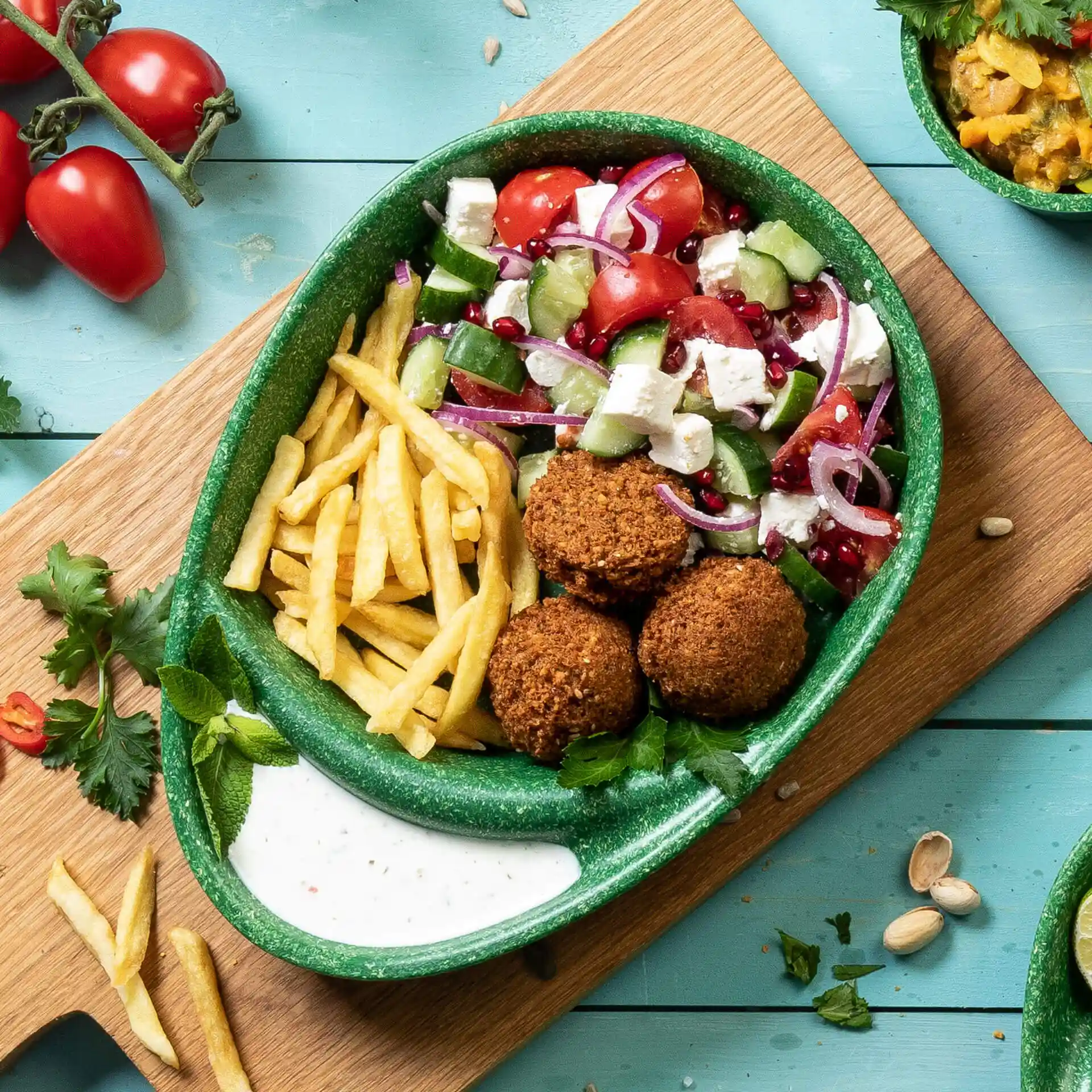 Assiette verte divisée avec des boulettes de falafel, des frites, une salade et de la sauce, placée sur une surface en bois avec des tomates et des herbes à proximité.