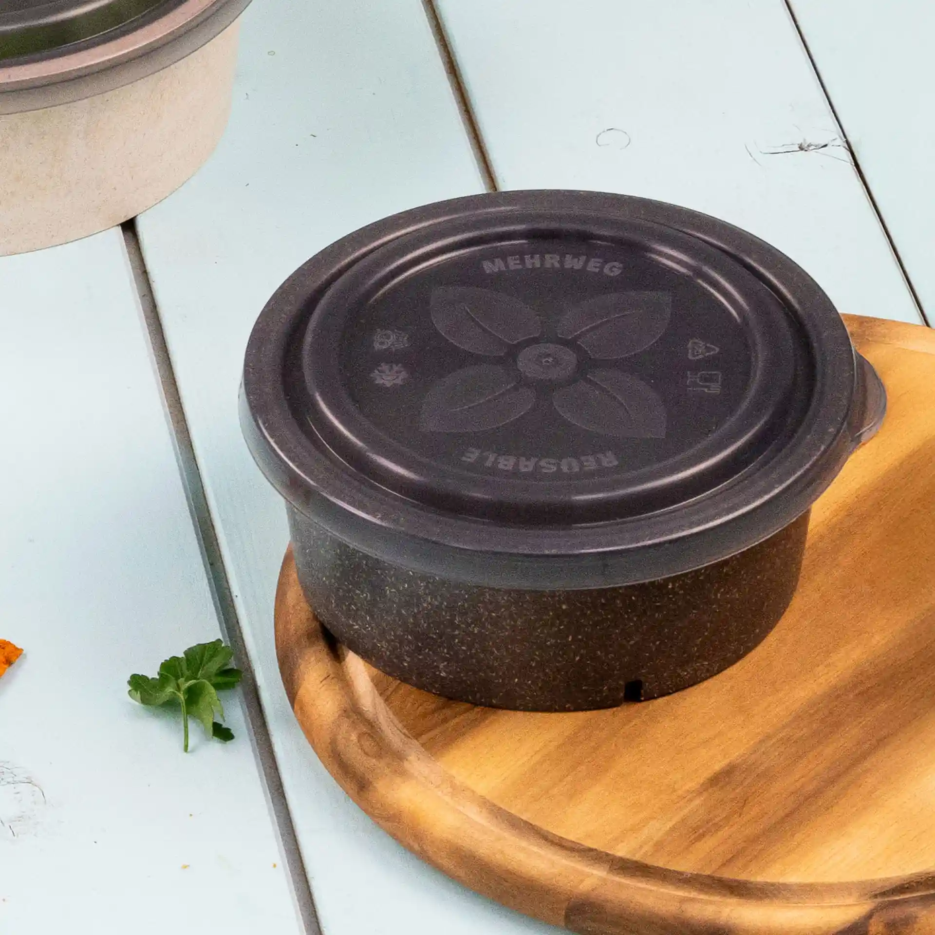 Bol rond gris réutilisable avec un couvercle noir, posé sur une planche en bois, avec un motif floral sur le couvercle.