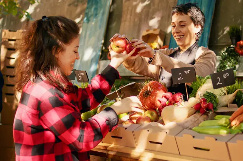 Femme proposant des pommes à un client à un stand de marché rempli de fruits et légumes variés.