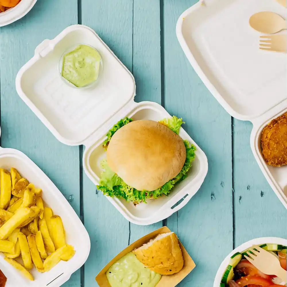 Un hamburger simple sur de la laitue dans un contenant à emporter blanc, avec des frites, une sauce et d'autres aliments visibles.