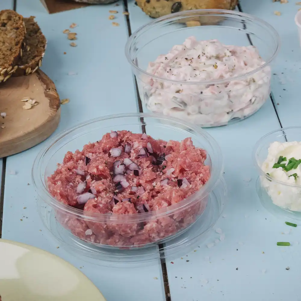 Viande crue hachée avec des oignons en dés dans un récipient transparent; à côté, une sauce crémeuse dans un autre récipient et un petit bol de fromage.