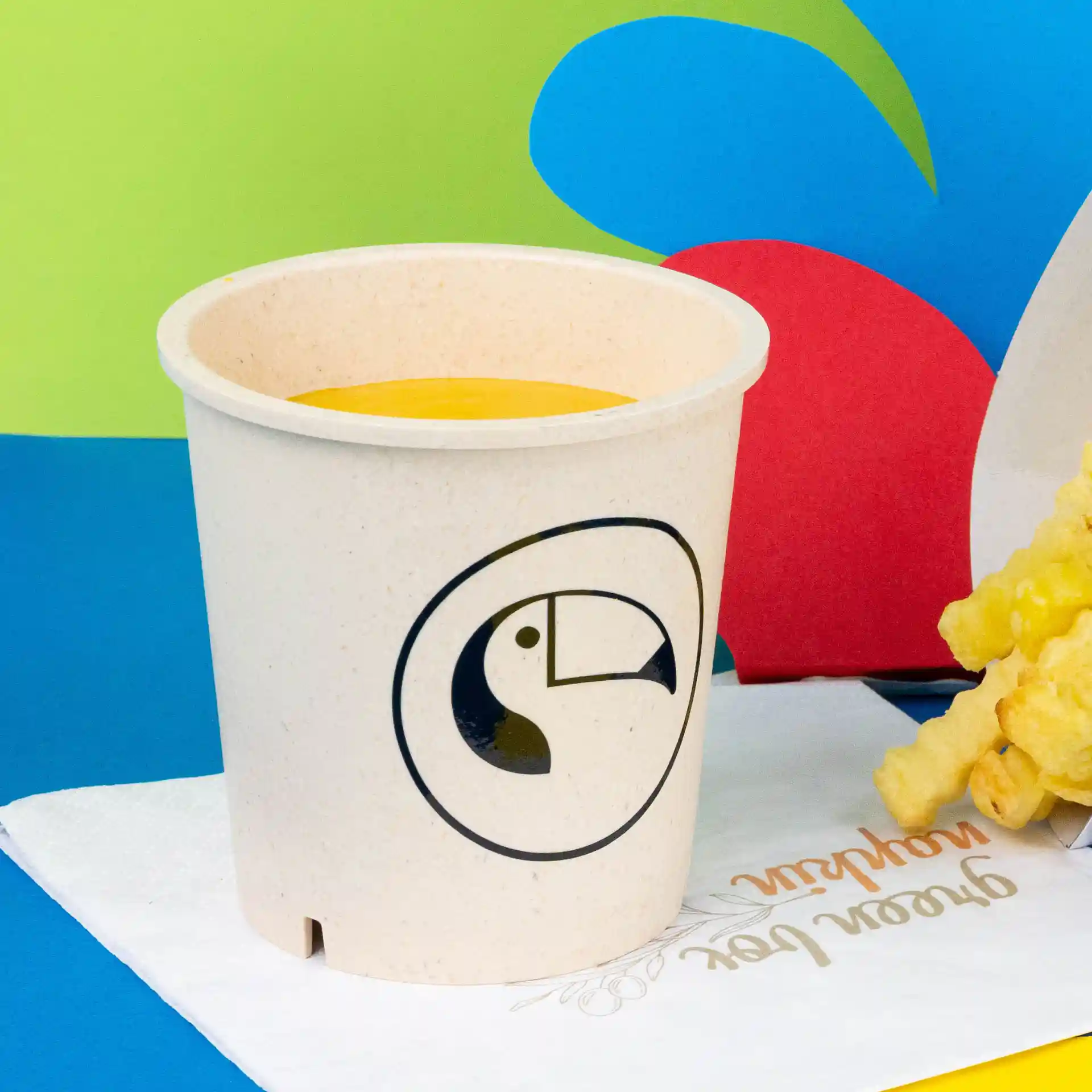 Bol rond crème réutilisable avec un logo de toucan, rempli de soupe jaune, posé sur une serviette.