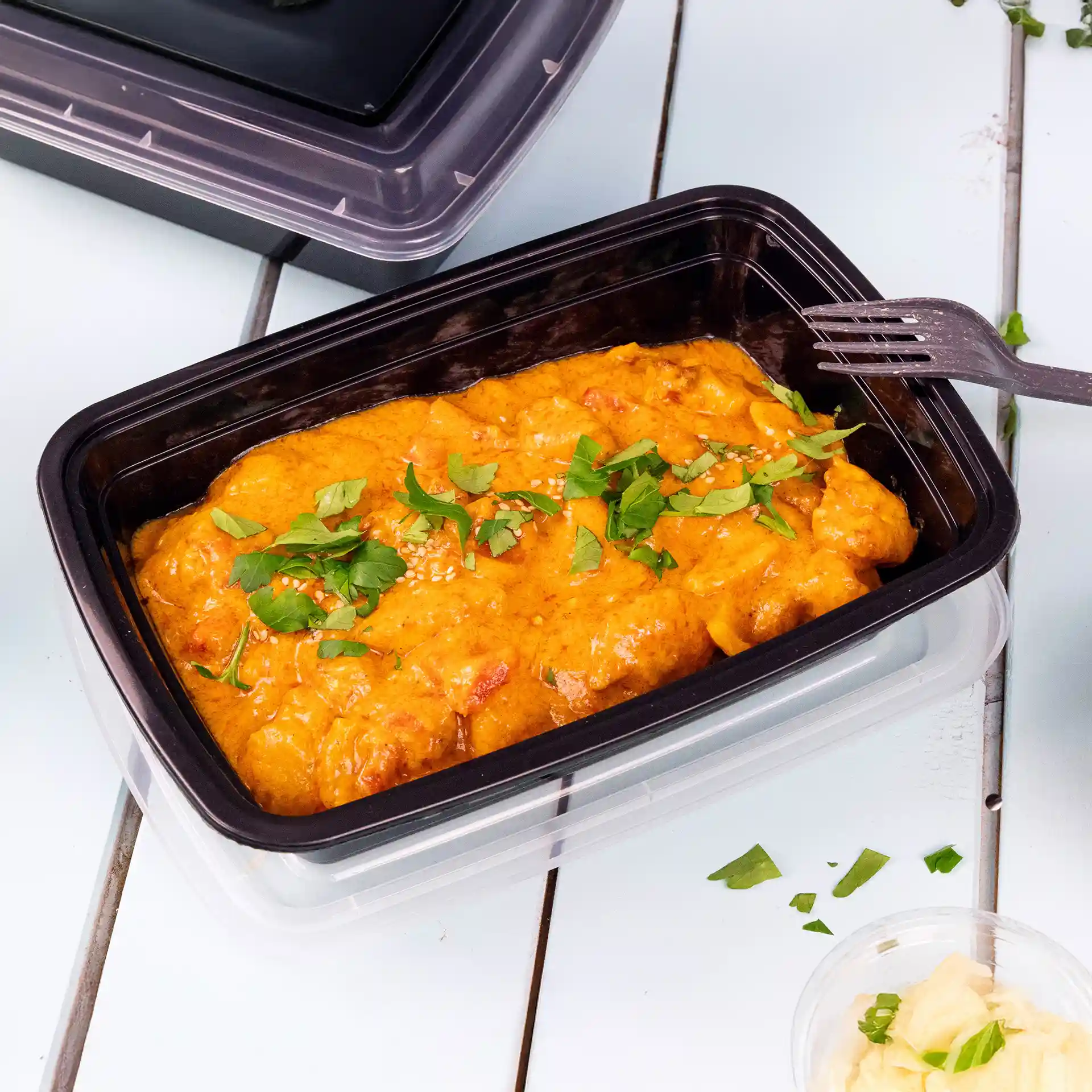 Conteneur rectangulaire noir réutilisable avec un couvercle, contenant des aliments orange garnis d'herbes vertes et de graines de sésame.