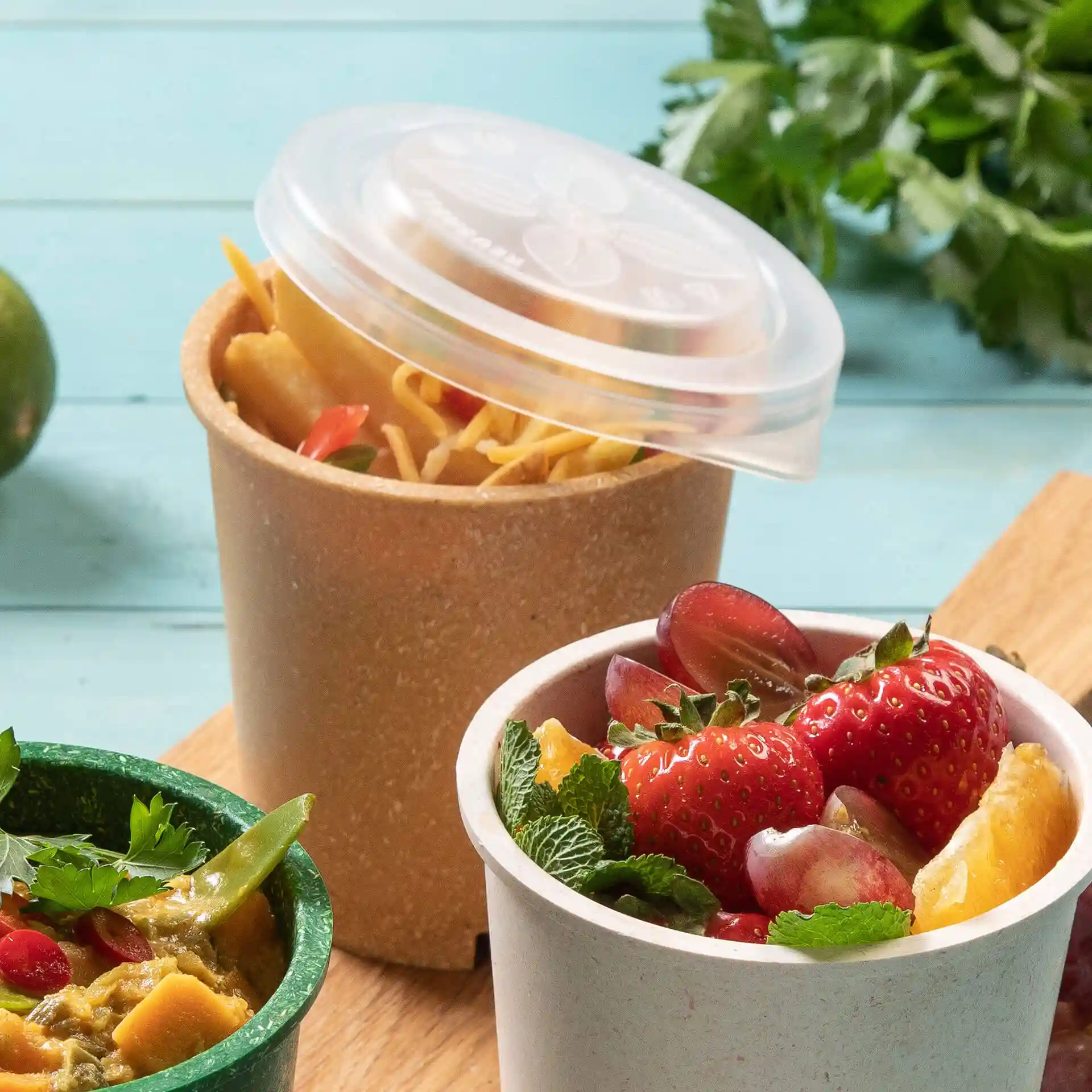 Image de deux tasses rondes, l'une avec un couvercle transparent contenant des nouilles, l'autre avec des fruits, sur une surface en bois.
