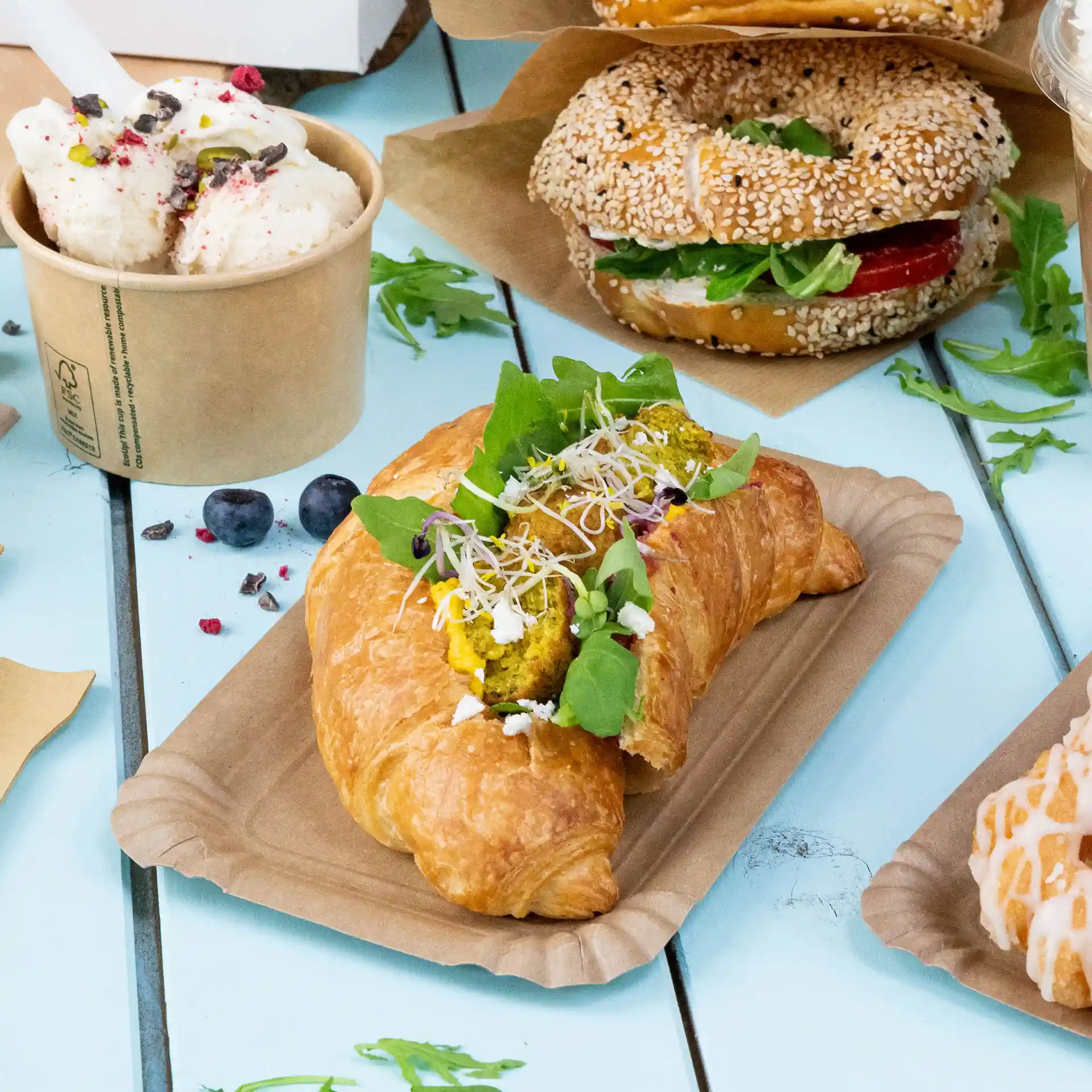 Croissant garni de feuilles vertes, de pousses et de garniture jaune sur une assiette en papier brun, avec d'autres aliments en arrière-plan.