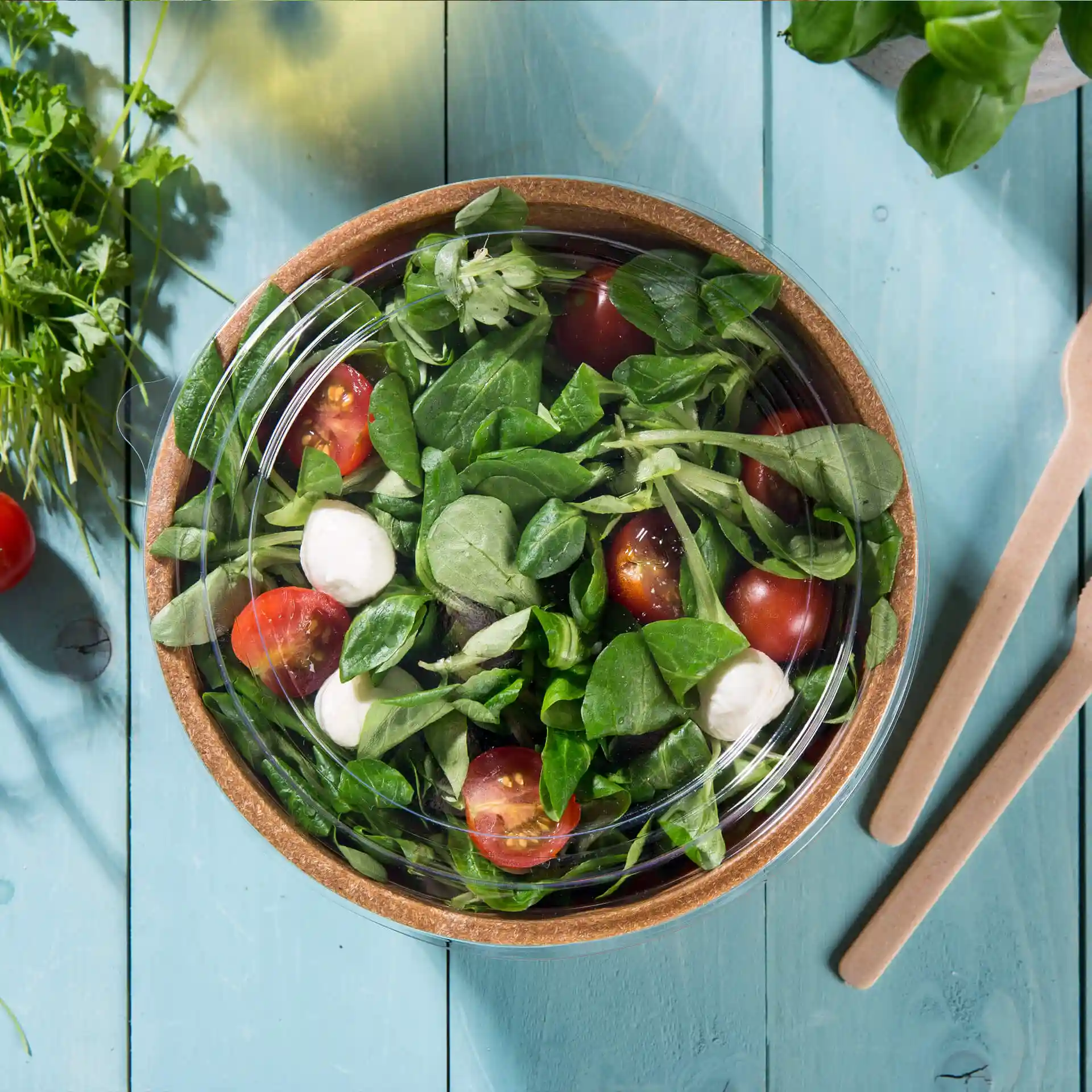 Couvercle rond transparent en PLA, placé sur un bol de salade avec des tomates et des épinards sur une surface en bois bleu.