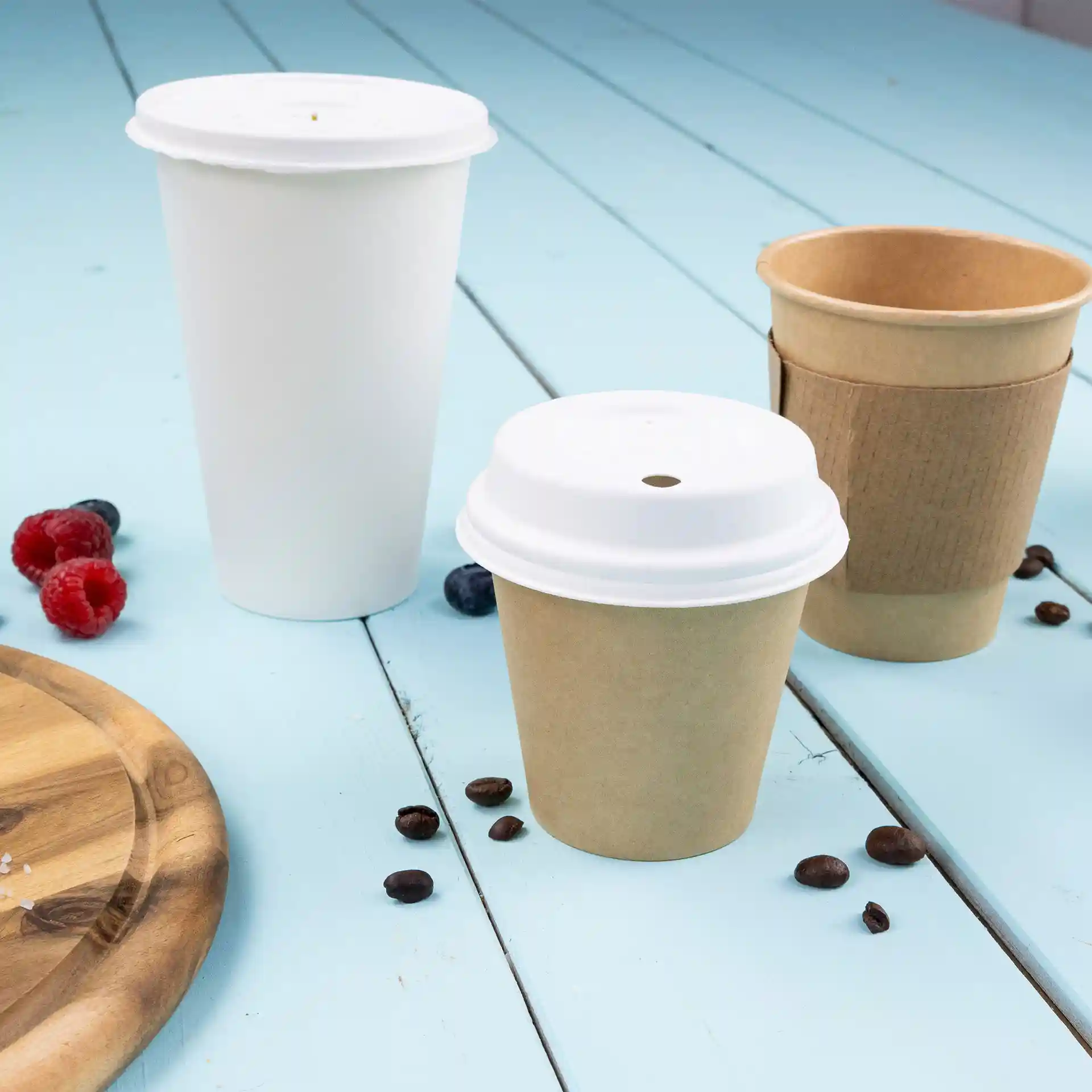 Image de trois gobelets en papier : un blanc, un marron avec un couvercle blanc, et un marron sans couvercle, sur une surface bleue avec des grains de café et des baies.