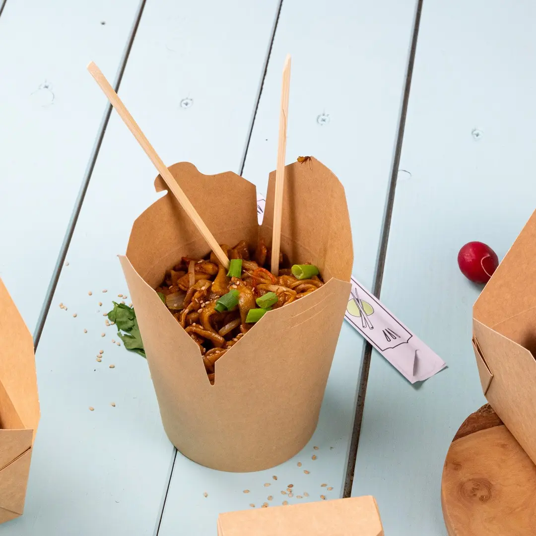 Boîte à emporter remplie de nouilles, garnie de poivrons verts et de graines de sésame, avec des baguettes insérées, sur une surface bleu clair.