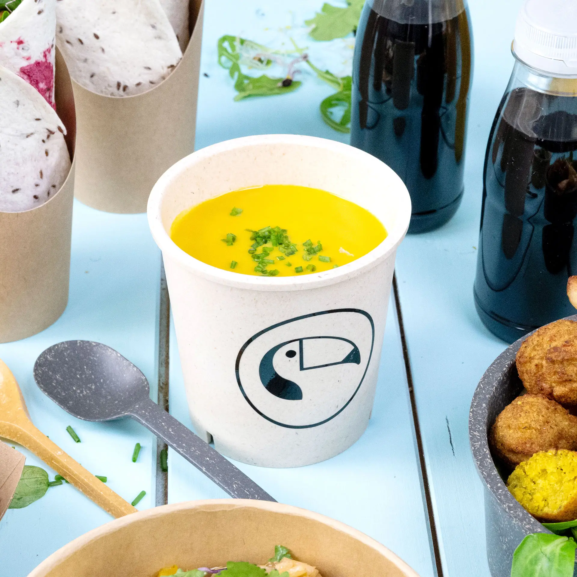 Gobelet à soupe réutilisable crème rempli de soupe jaune et garni de ciboulette, sur un fond bleu avec d'autres aliments.