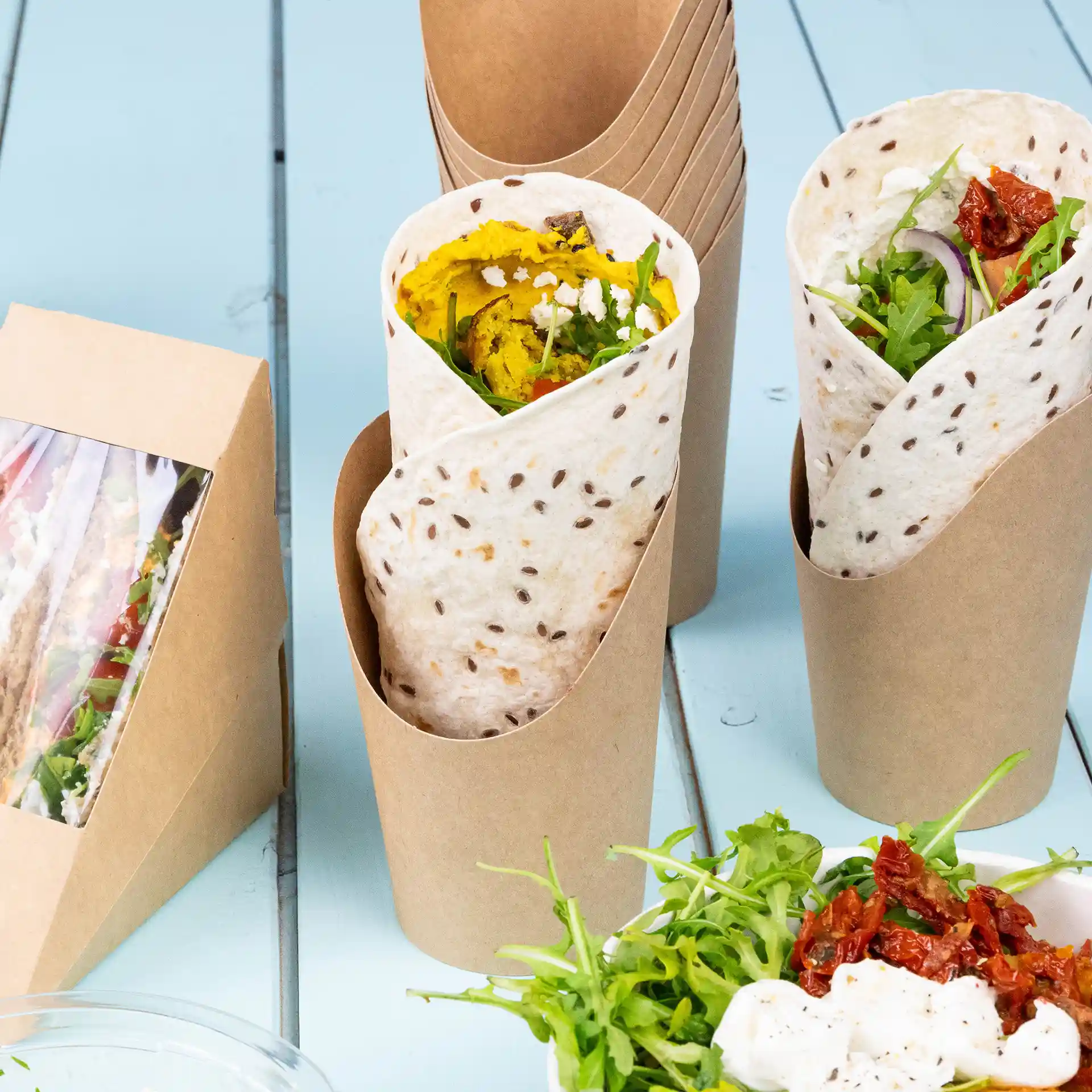 Image montrant deux gobelets en carton brun, l'un rempli de nourriture et une salade d'accompagnement avec des légumes verts et des tomates séchées.