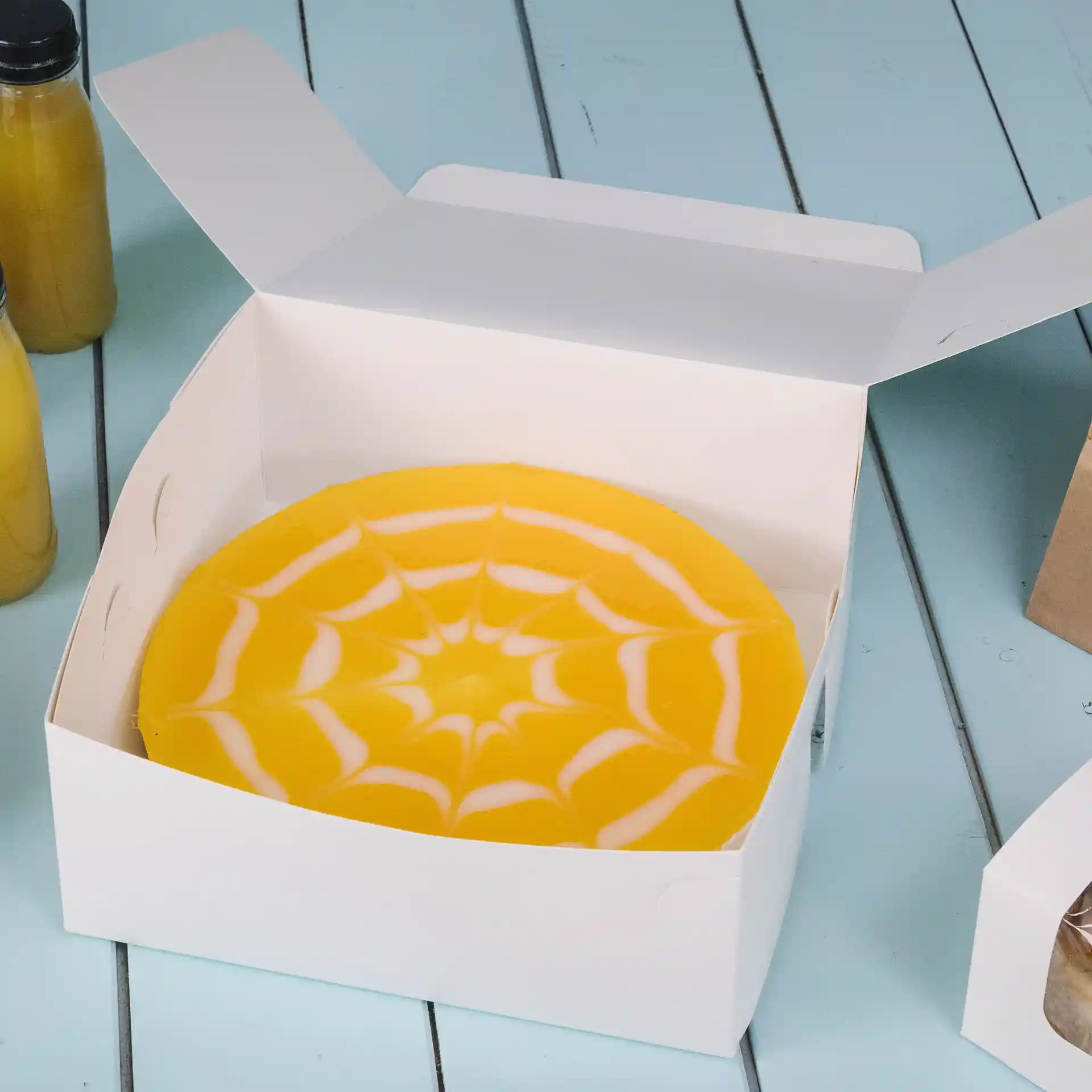 Boîte à gâteau blanche avec un gâteau jaune présentant un design de toile d'araignée à l'intérieur, placée sur une surface bleue.