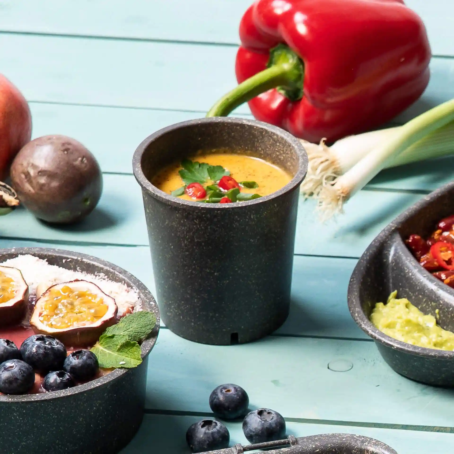Gobelet réutilisable gris avec de la soupe, garni d'herbes et de piment, entouré de fruits et légumes sur une surface bleue.