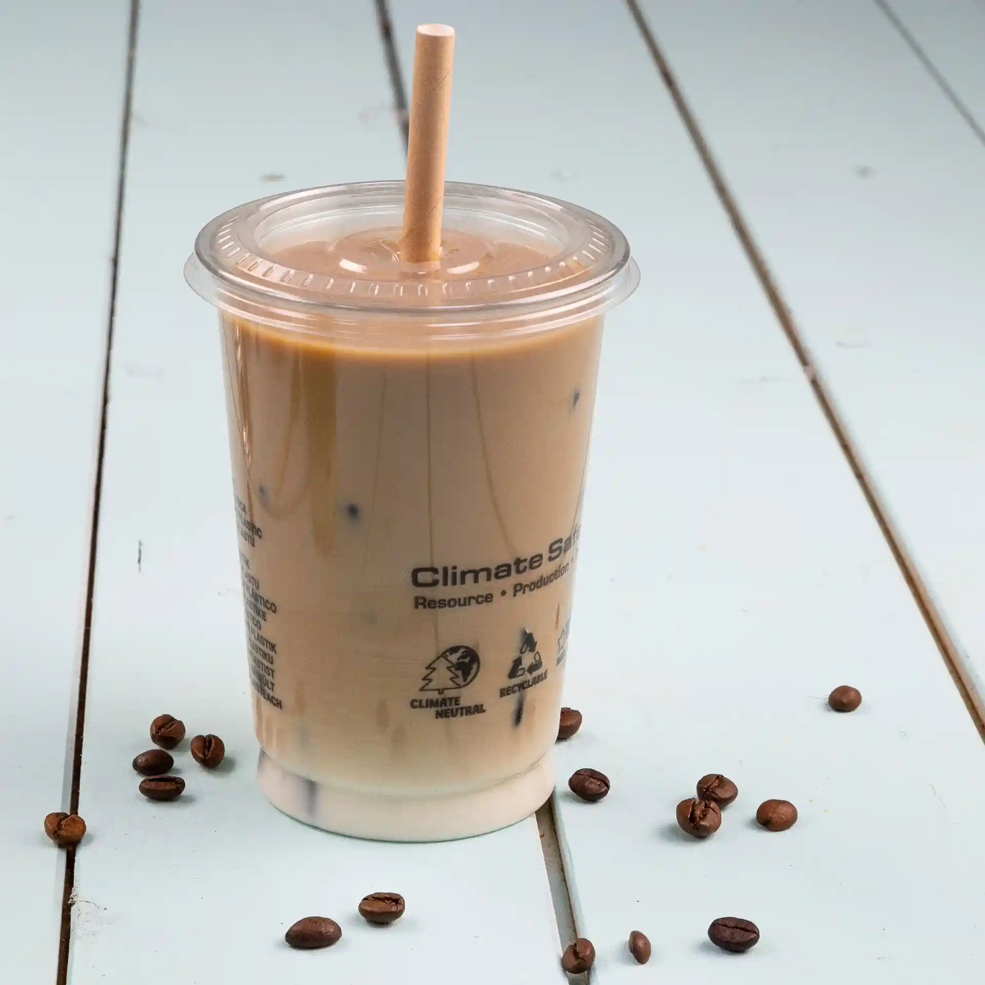 Gobelet transparent avec café glacé, surmonté d'une paille, posé sur une surface bleu clair avec des grains de café éparpillés.