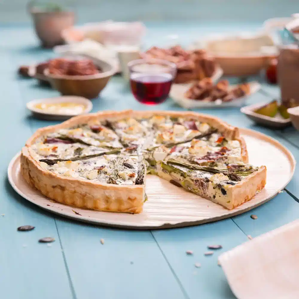 Quiche savoureuse sur une assiette en bois, avec une part retirée, entourée de divers plats et d'un verre de boisson rouge.