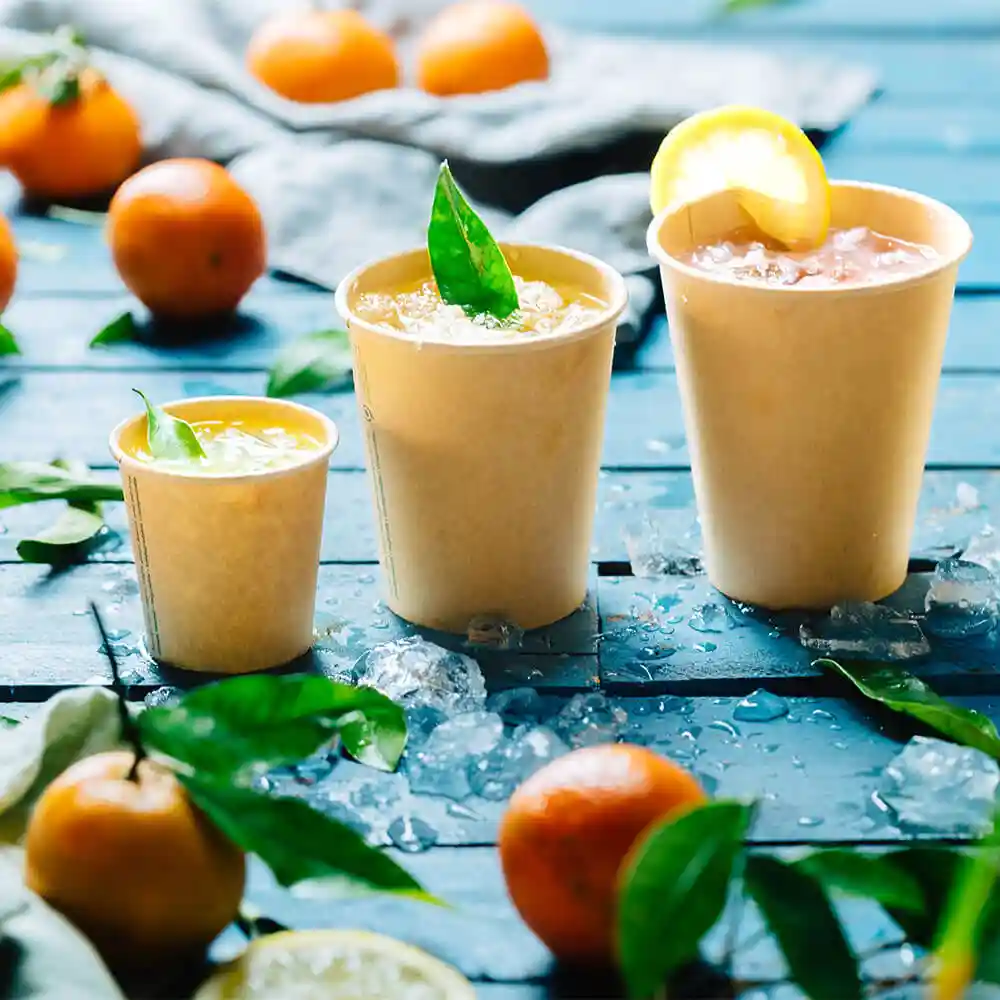 Trois gobelets en papier marron remplis de boissons, placés sur une surface en bois bleu avec de la glace et des agrumes.