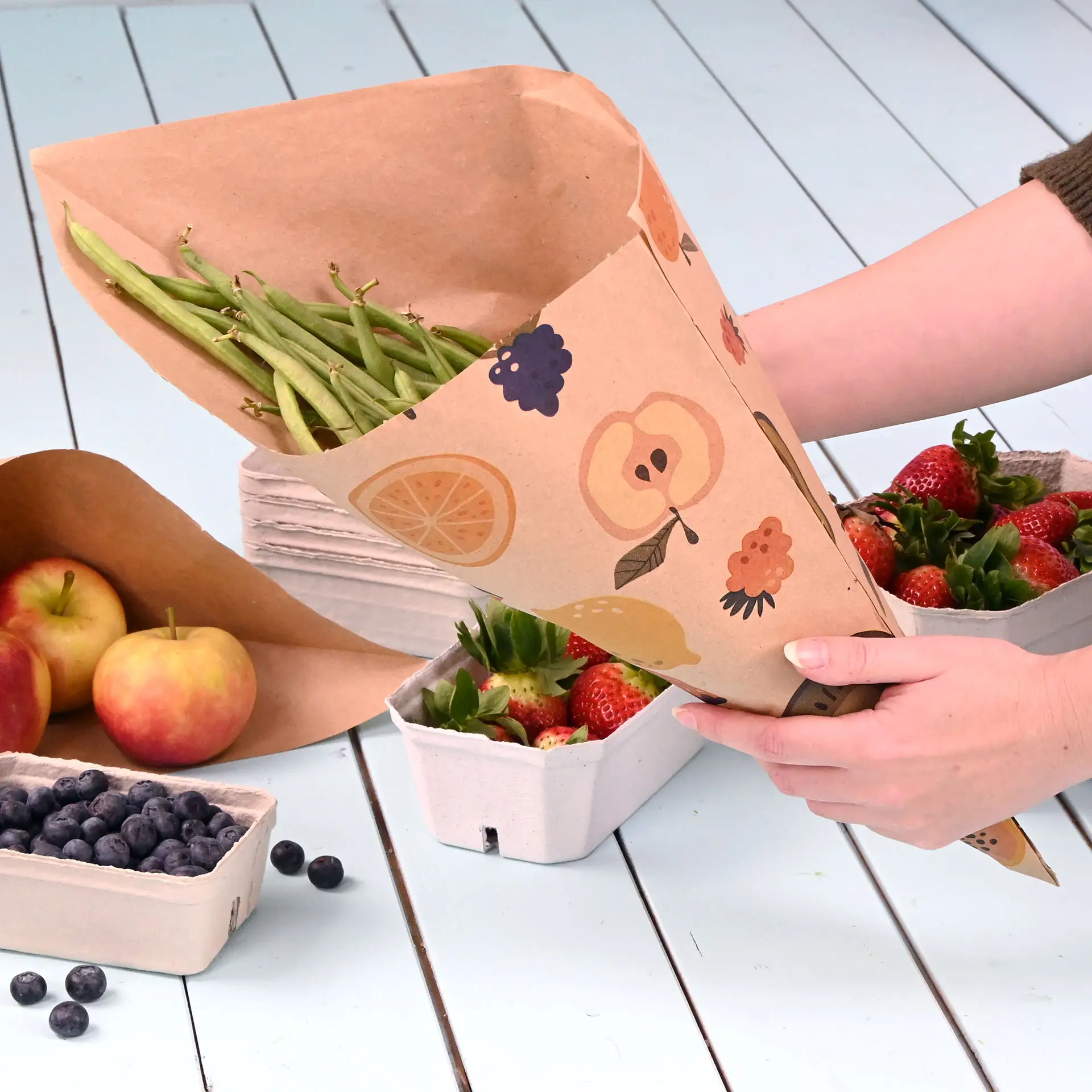 Une main tient un cône en papier brun rempli de haricots verts, entouré de fraises, pommes et myrtilles sur une surface bleu clair.